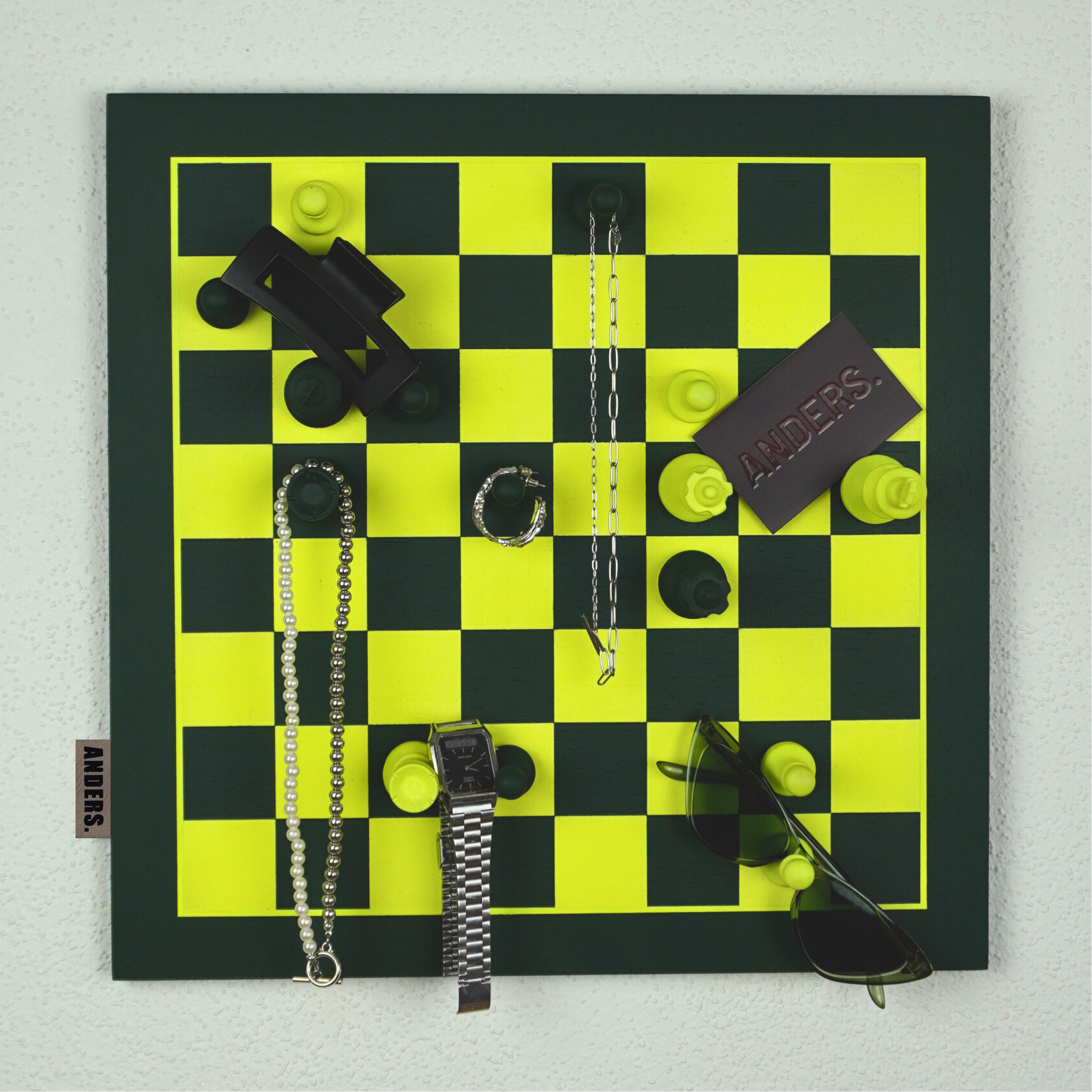 Ein Schachbrett mit schwarzen und gelben Figuren, umgeben von verschiedenen Gegenständen wie Schmuck, Uhren, Sonnenbrille, und einem Zettel mit dem Schriftzug "ANDERS".