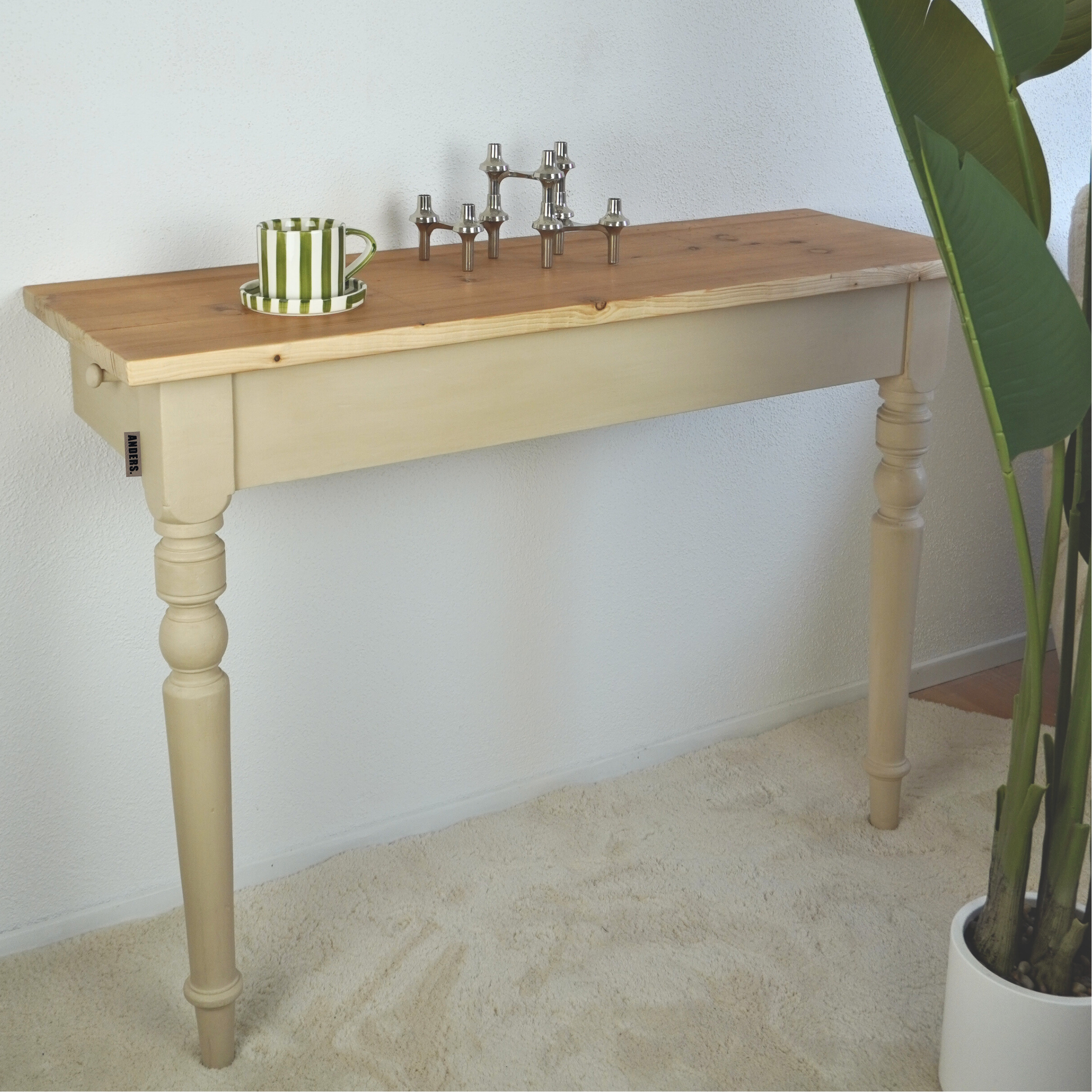 Ein improvisiertes Waschbecken mit einem Holztisch, einer Wasserhahn-Armatur und einem grünen und weißen gestrichenen Becher mit Untertasse. Es steht an einer weißen Wand und neben einem großen Zimmerpflanzenkübel.