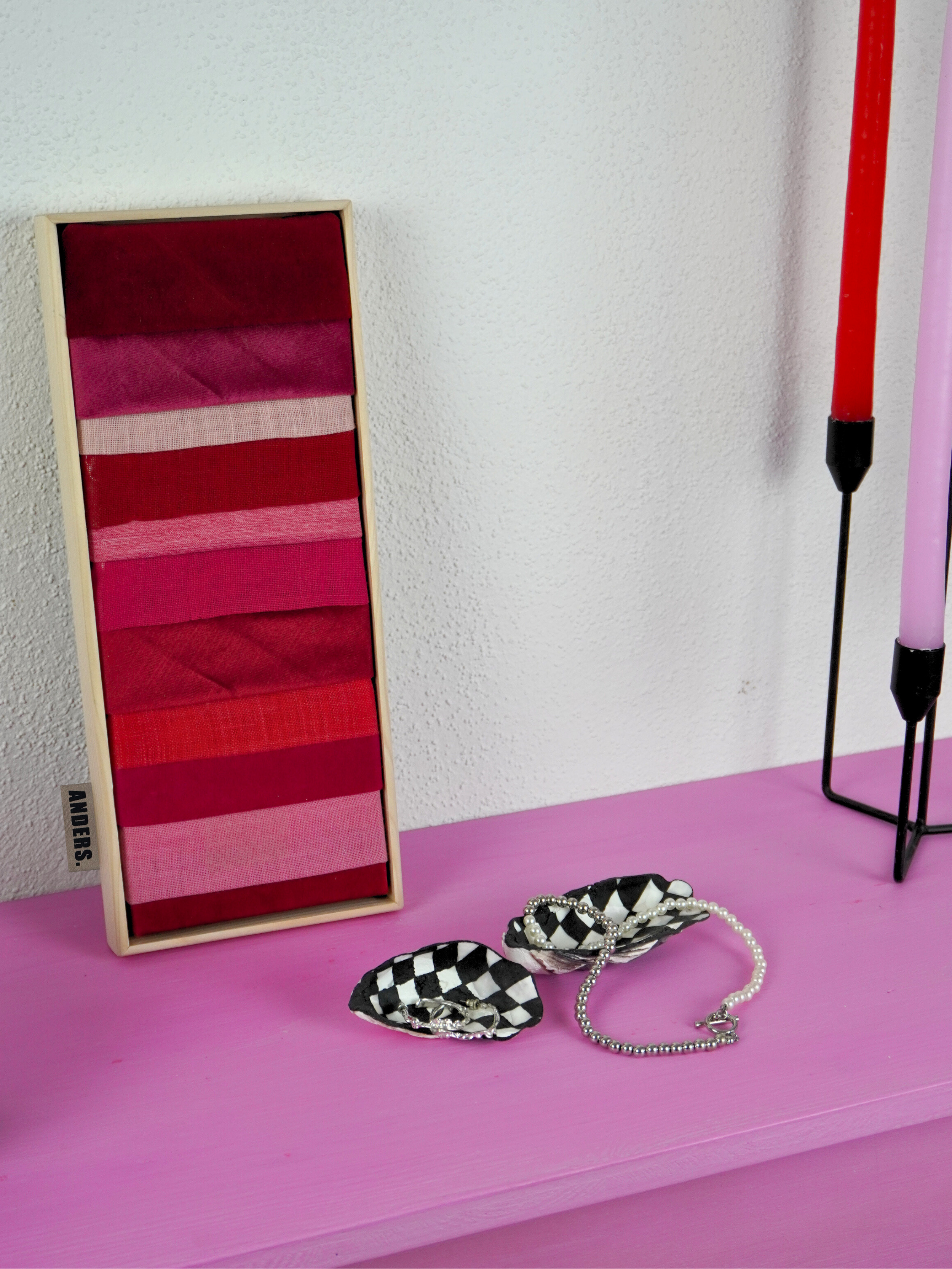 Ein geöffnetes Farbband-Set mit verschiedenen Rottönen auf einer pinken Oberfläche, daneben eine schwarze und weiße Checkered-Skull-Form mit Schmuck, und zwei hohe Kerzen, eine rot und eine pink.