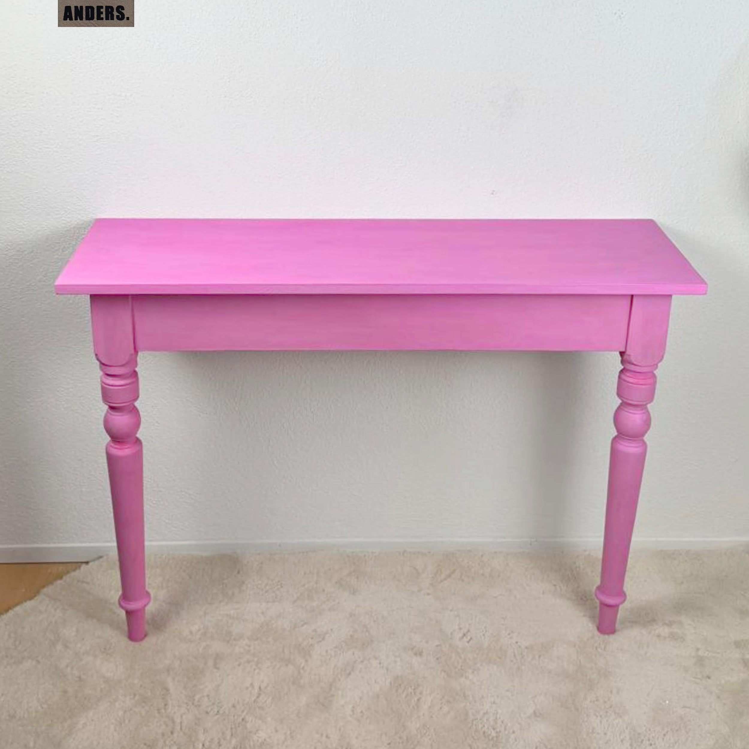 Ein rosa Holzkonsole mit gedrechselten Beinen steht vor einer weißen Wand, oben links ein schwarzes und braunes Etikett mit dem Wort 'ANDERS.'.