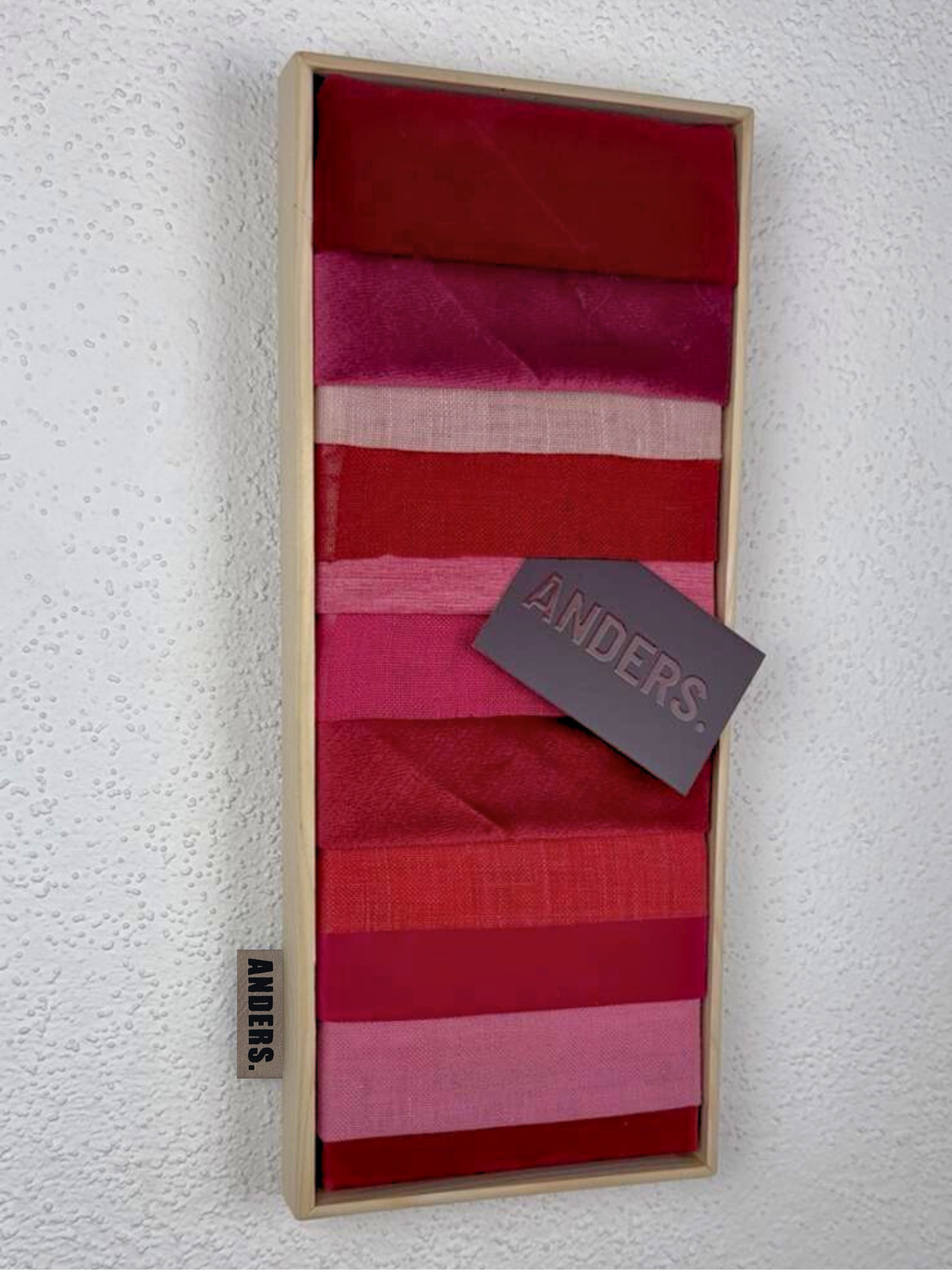 Ein Holzrahmen mit verschiedenen roten und pinkfarbenen Textilproben, eine Karte mit der Aufschrift 'ANDERS.' liegt darauf.