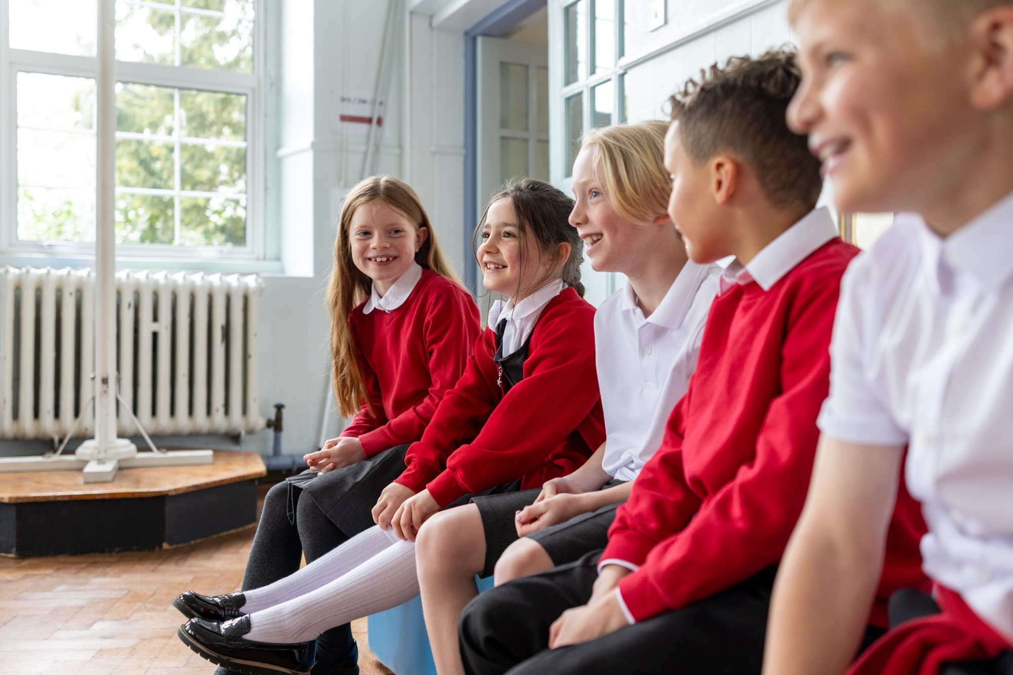 Group of school children sitting on a bench, smiling and talking in a classroom with large windows and a radiator.