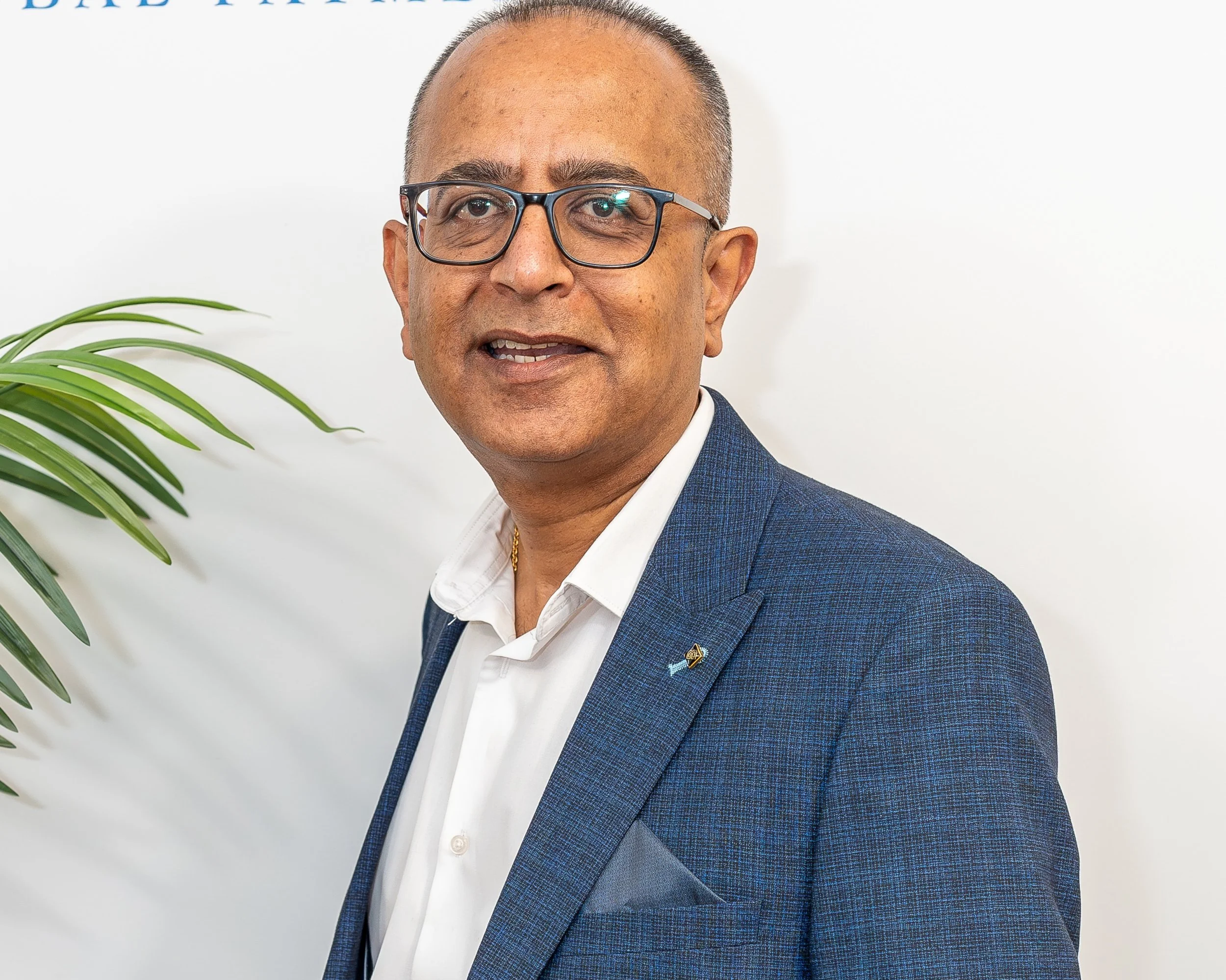 A man wearing glasses and a navy blue suit with a white shirt, smiling, standing next to a potted plant with green leaves, against a white wall background.
