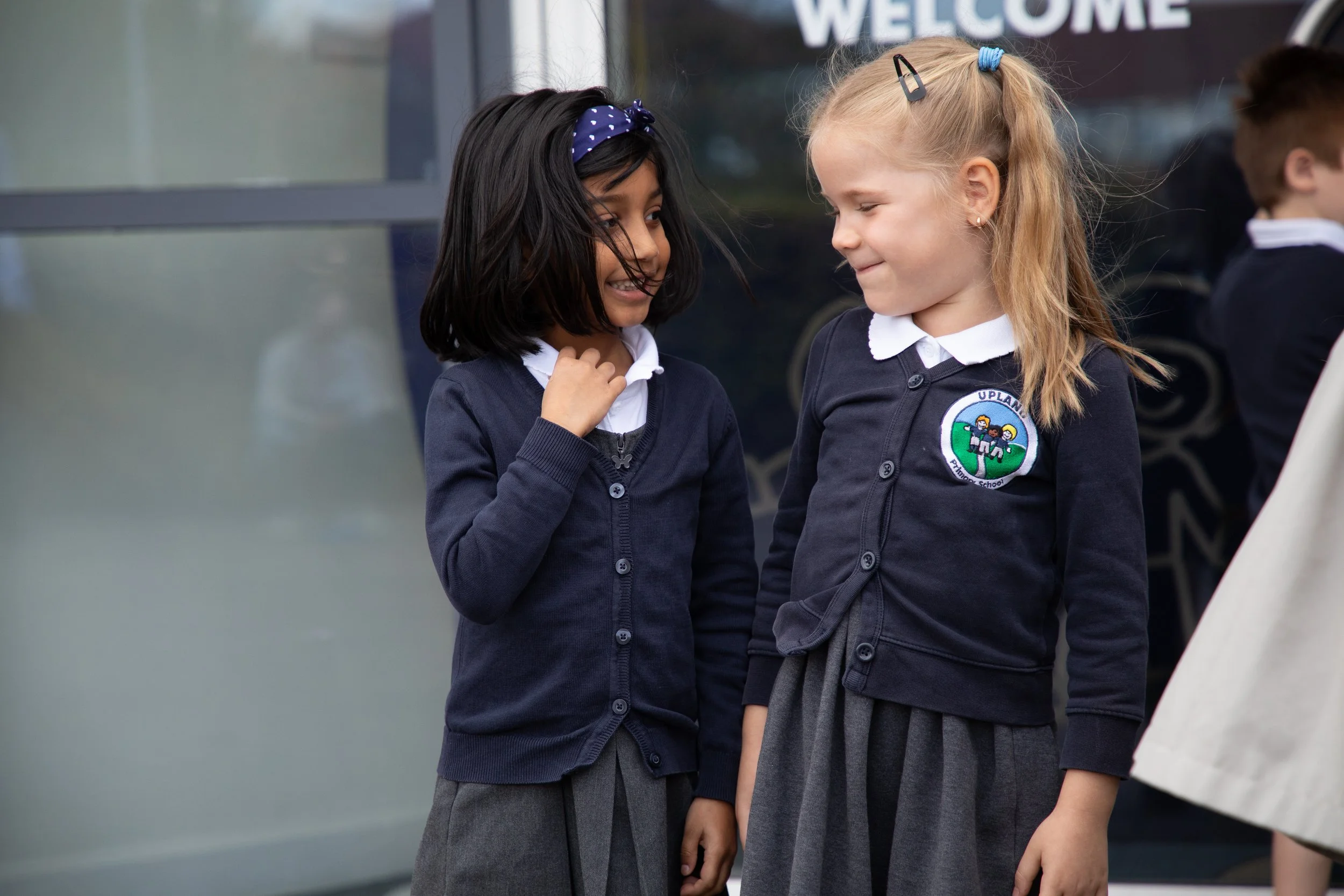 Two young girls in school uniforms smiling and talking outside, with a school building and other children in the background.