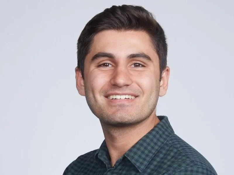 A young man with dark, short hair and a light beard, smiling, wearing a dark checkered shirt, against a plain light gray background.