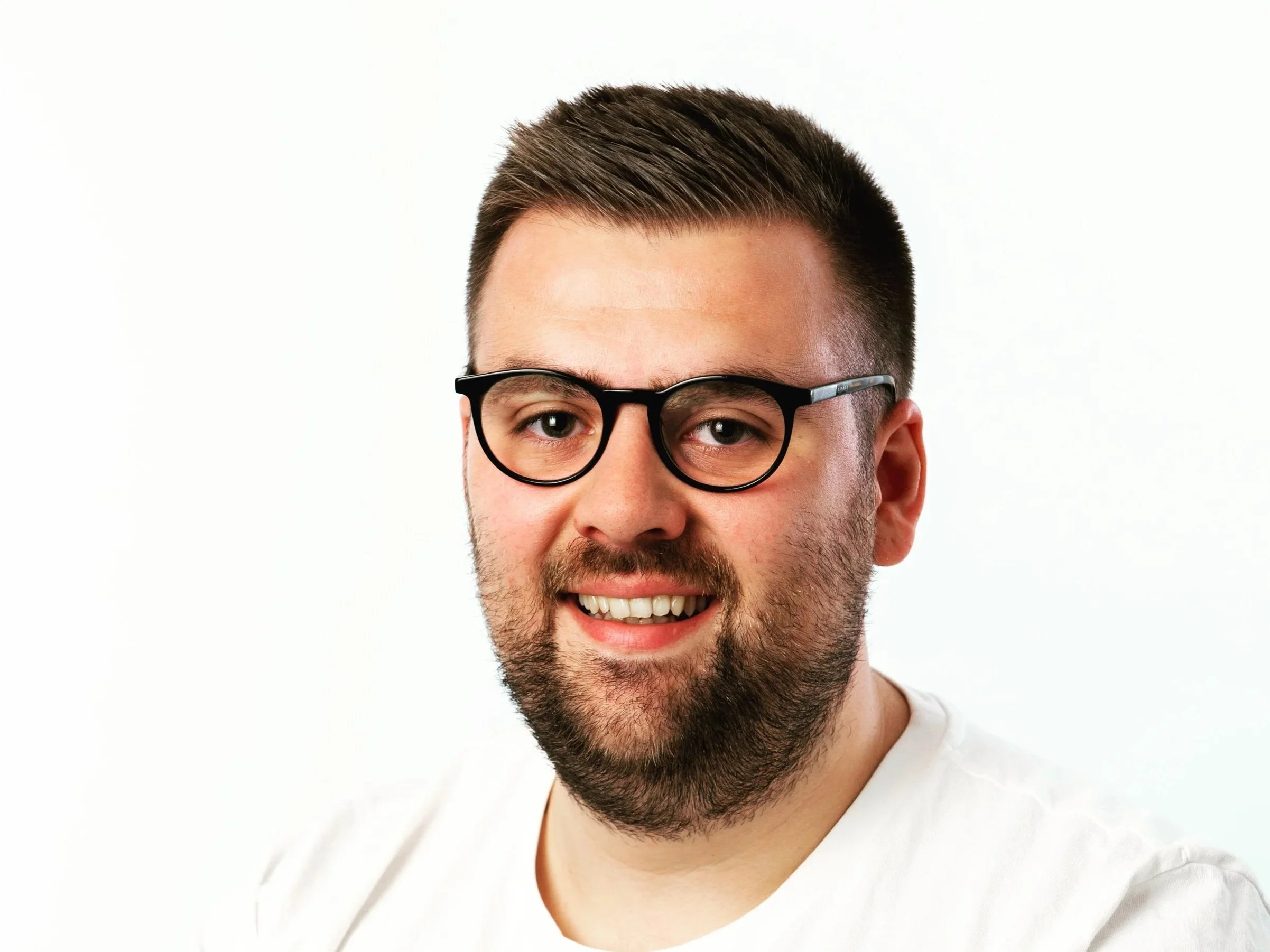 Close-up portrait of a young man with short brown hair, beard, wearing glasses and a white shirt, smiling against a plain white background.
