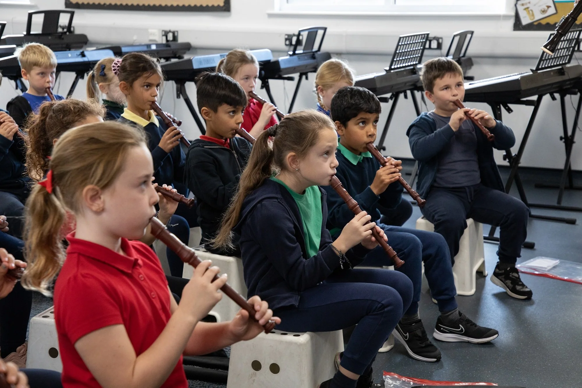 Group of young children sitting on white plastic stools, playing recorders in a music classroom with keyboards in the background.