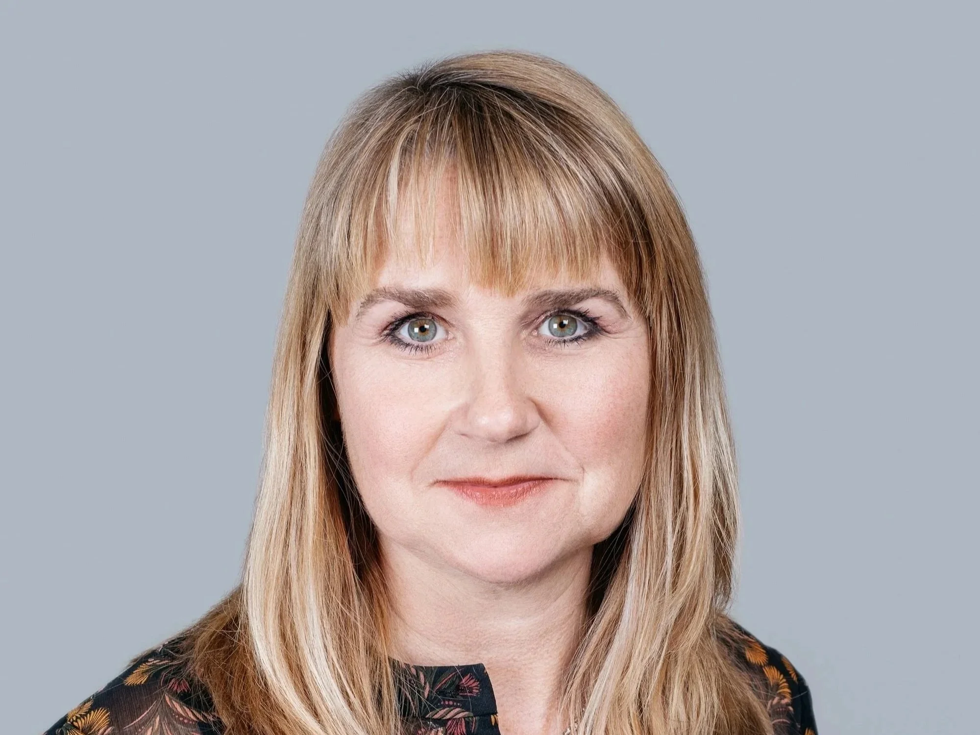 Headshot of a woman with blonde hair and blue eyes, wearing a dark patterned top, against a plain gray background.