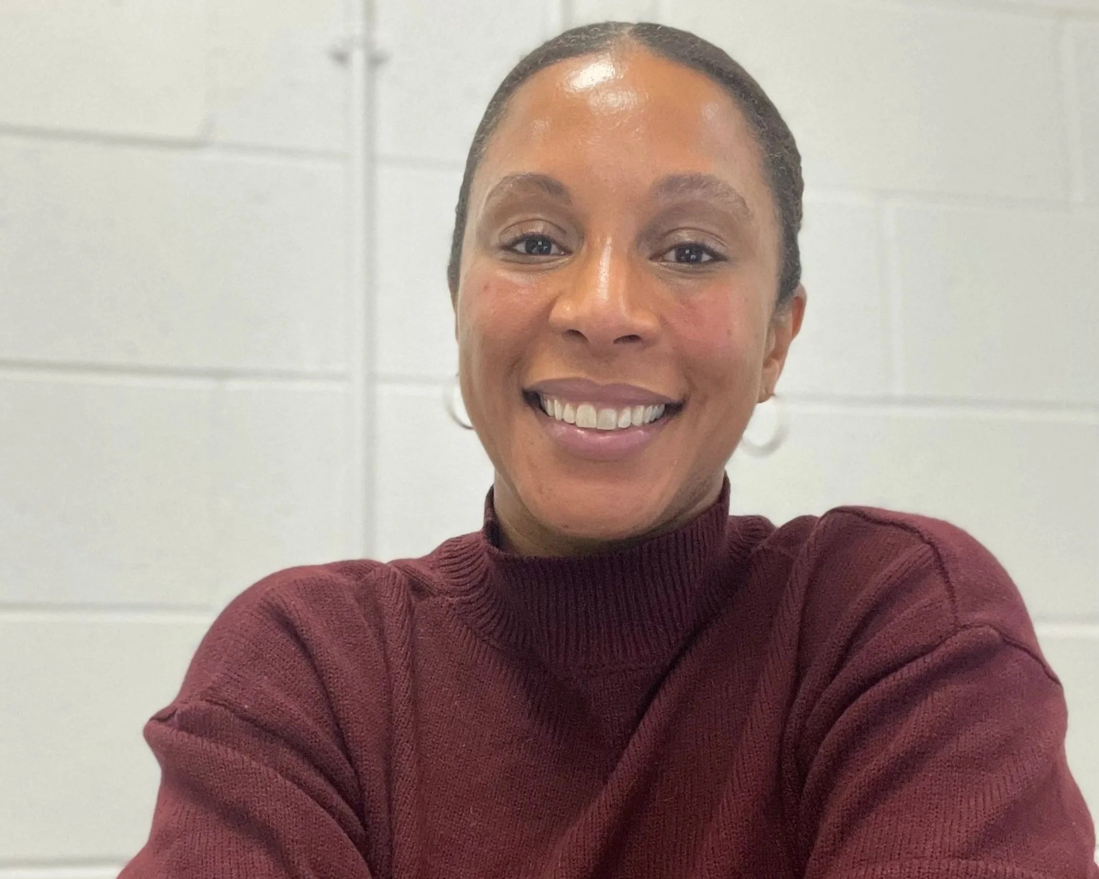 A smiling woman with dark hair pulled back, wearing a maroon sweater, posing indoors in front of a plain white wall.