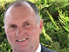 Close-up of a smiling man with short, gray hair outdoors with green foliage in the background.