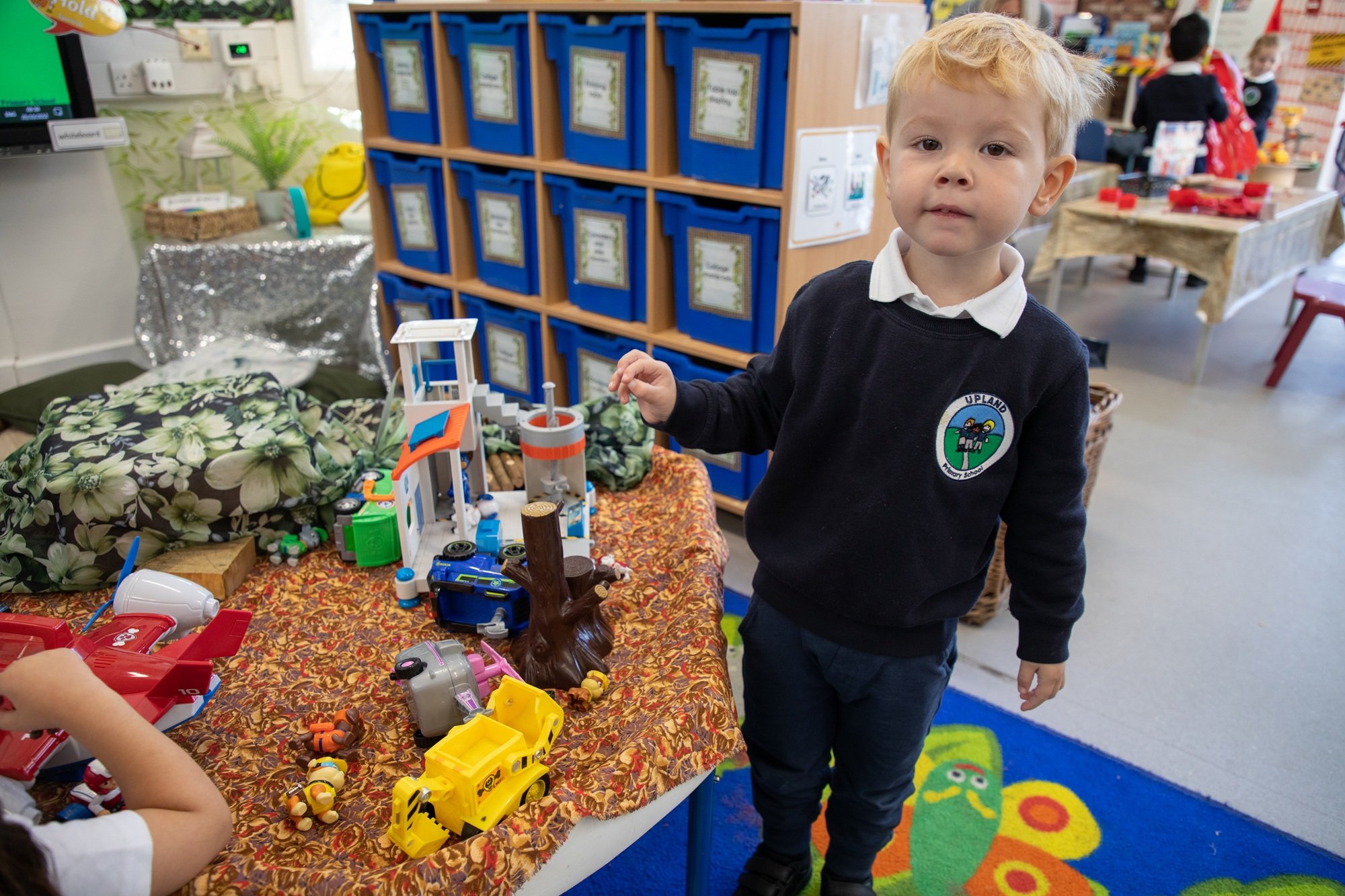 A young boy with blonde hair wearing a school uniform standing next to a table with toy vehicles, including a yellow truck, a red airplane, and other small toys, in a colorful classroom or play area.