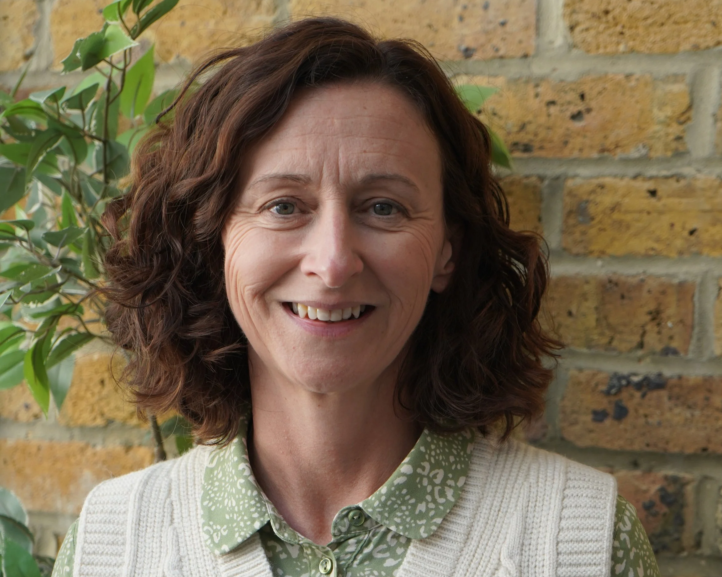 A woman with wavy brown hair smiling, wearing a green patterned shirt and cream-colored vest, standing against a brick wall with some green plants in the background.