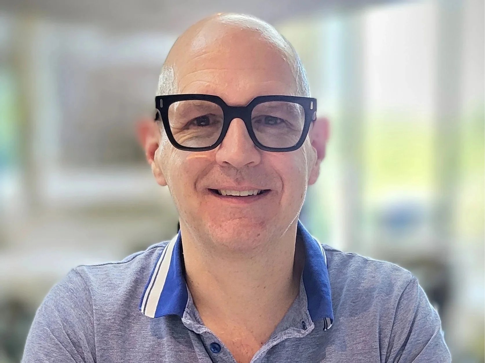 A smiling middle-aged man with a bald head, wearing large black glasses, in a casual gray polo shirt with a blue and white collar, indoors with blurred background.