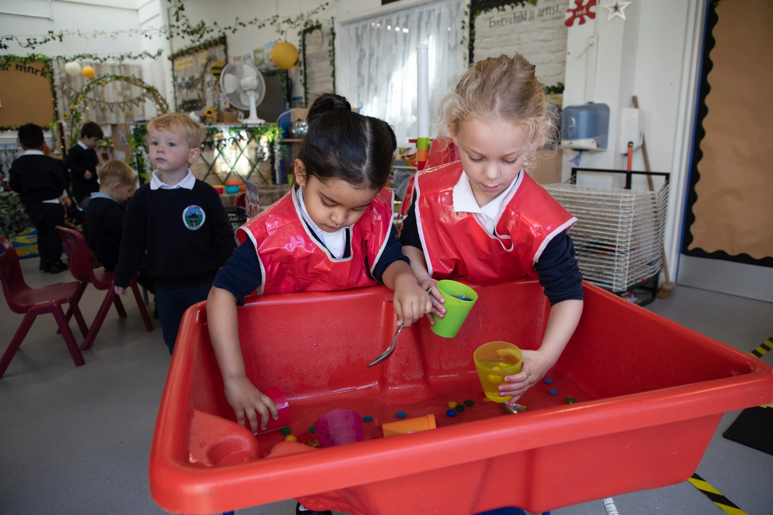Two young girls wearing red aprons playing a game with cups and small objects at a school or daycare. Other children in uniforms are seen in the background in a decorated classroom.