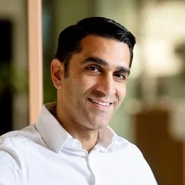 Headshot of a smiling man with dark hair, wearing a white shirt, in an indoor setting with blurred background.
