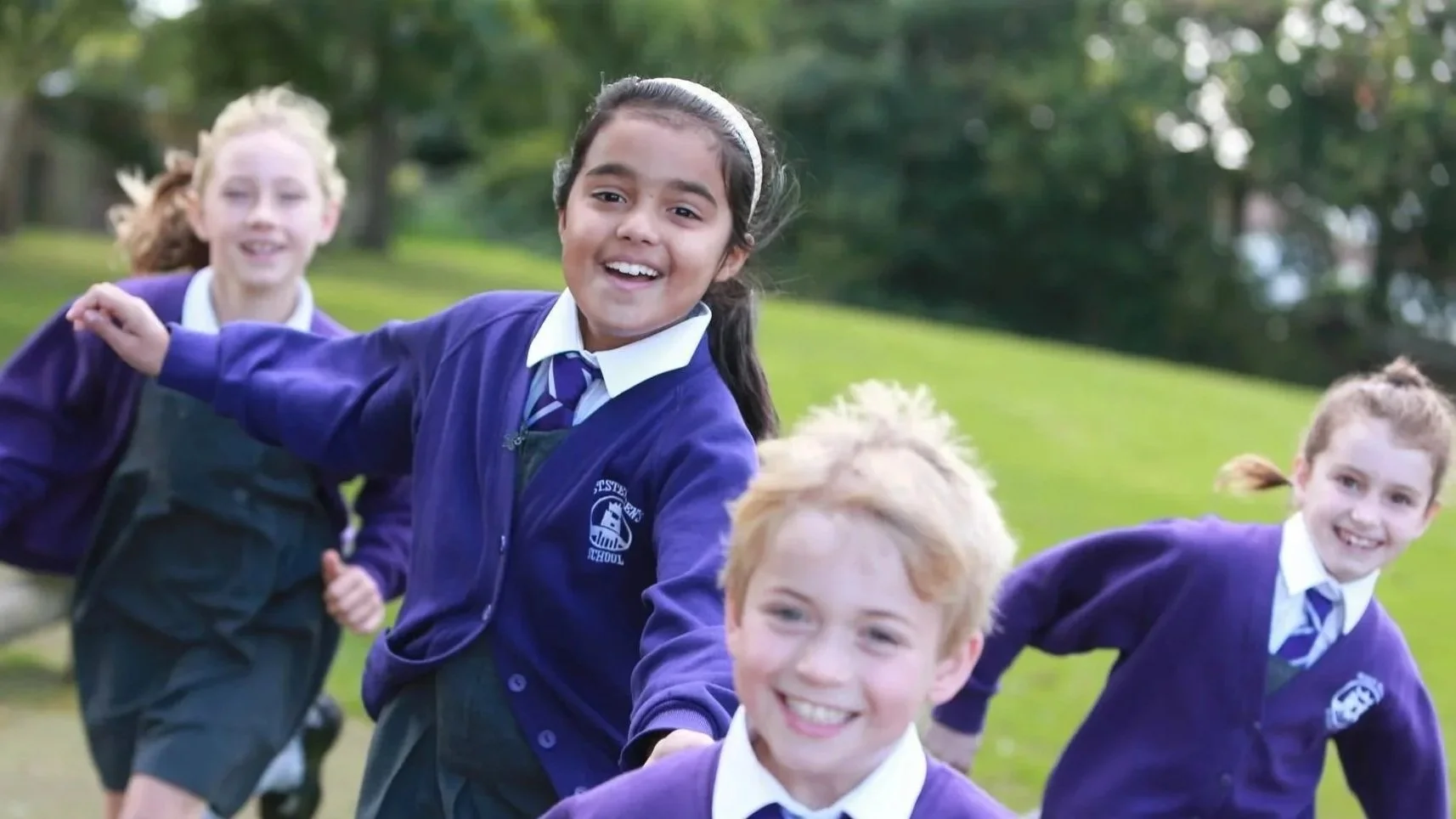 Four children in school uniforms playing outdoors on a grassy field, smiling and running.