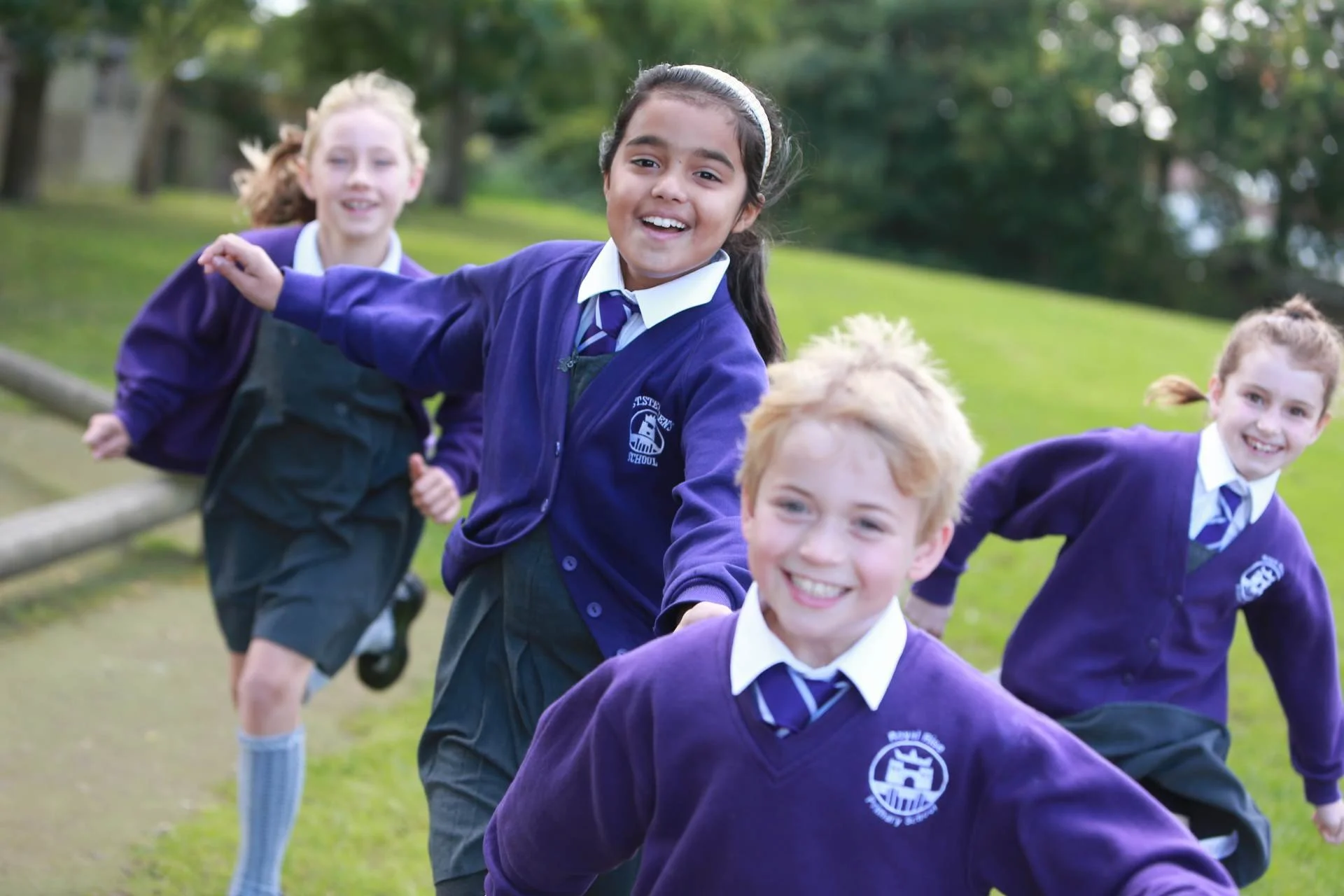 Four children in purple school uniforms running and smiling outdoors on a grassy area with trees in the background.