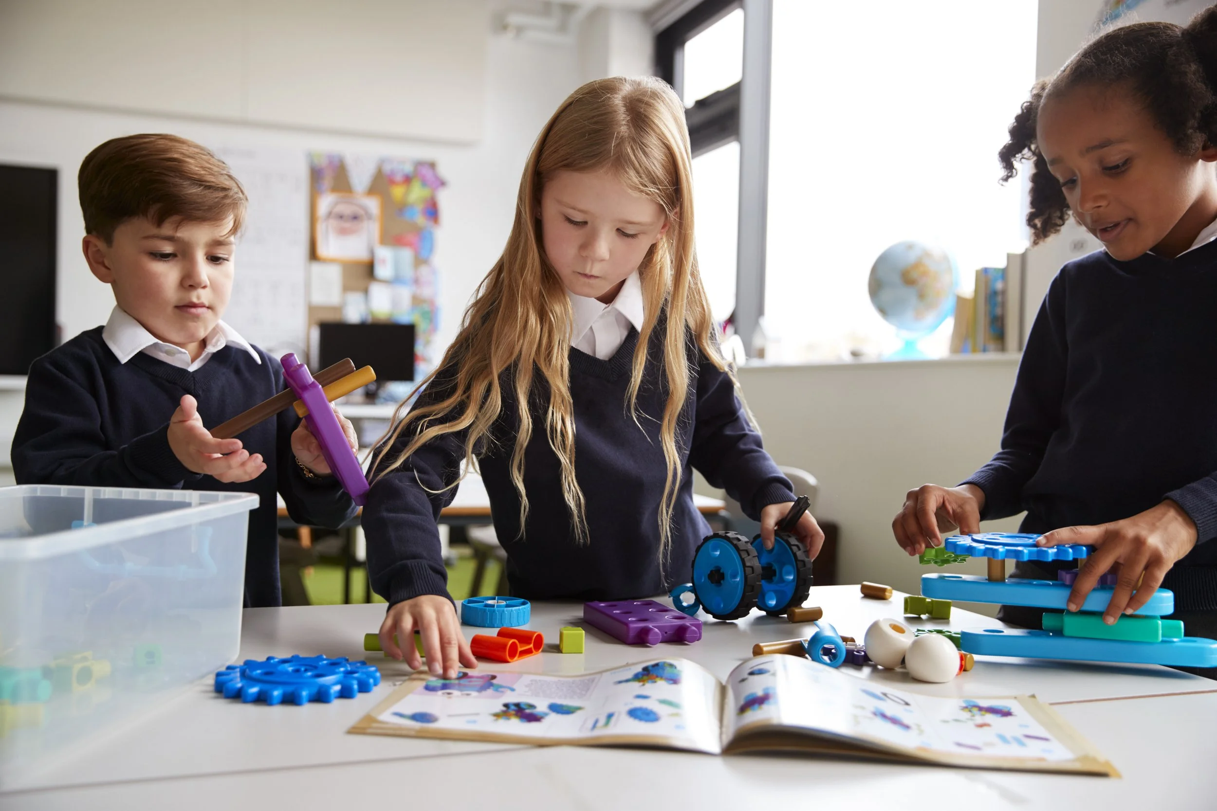 Three children in school uniforms building with colorful plastic construction toys at a table in a classroom.