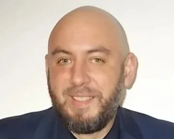 A man with a beard and bald head smiling, wearing a dark blazer against a light background.