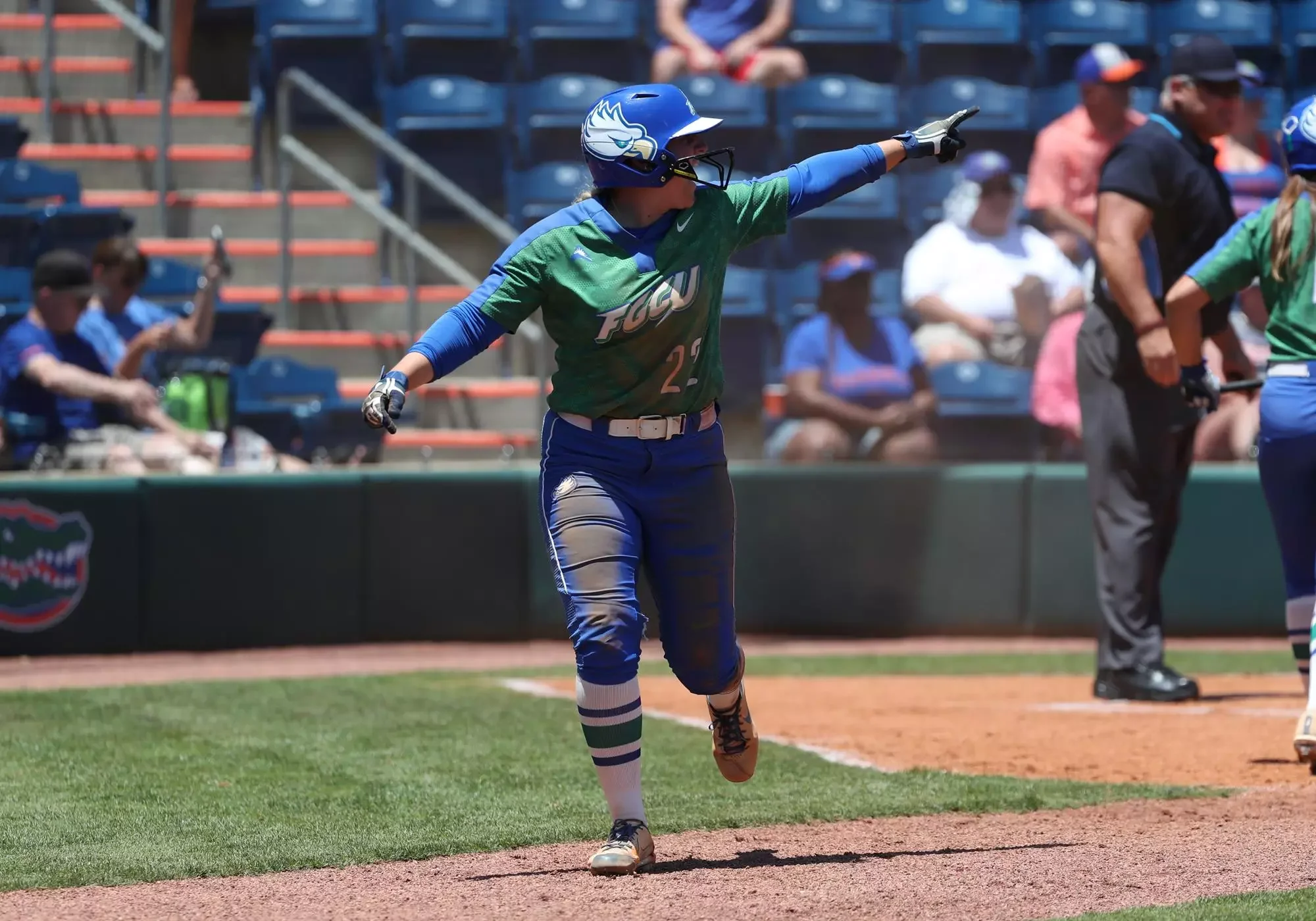 A female softball player in green and blue uniform celebrating on the field, pointing with her right arm after a play, with spectators in the background.