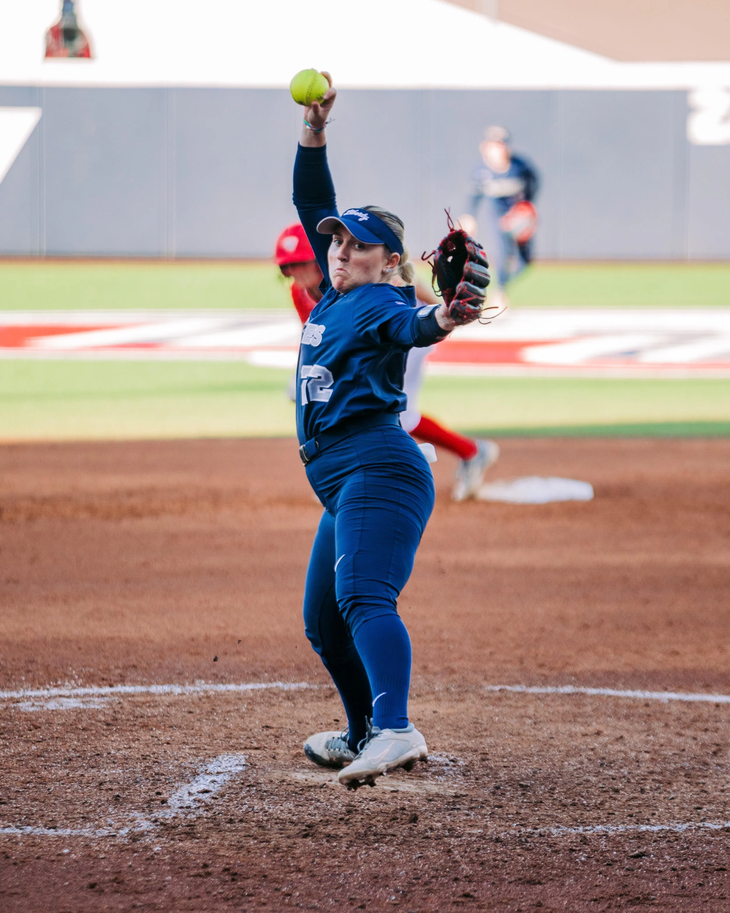Female softball pitcher in a blue uniform winding up to throw a pitch, with a yellow softball in her hand, on a dirt field.