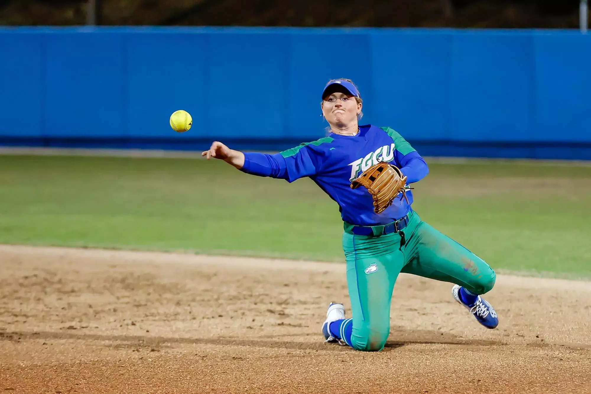 A female softball player in a blue and green uniform with a glove on her left hand is throwing a yellow softball while kneeling on the infield dirt, with a blue fence in the background.