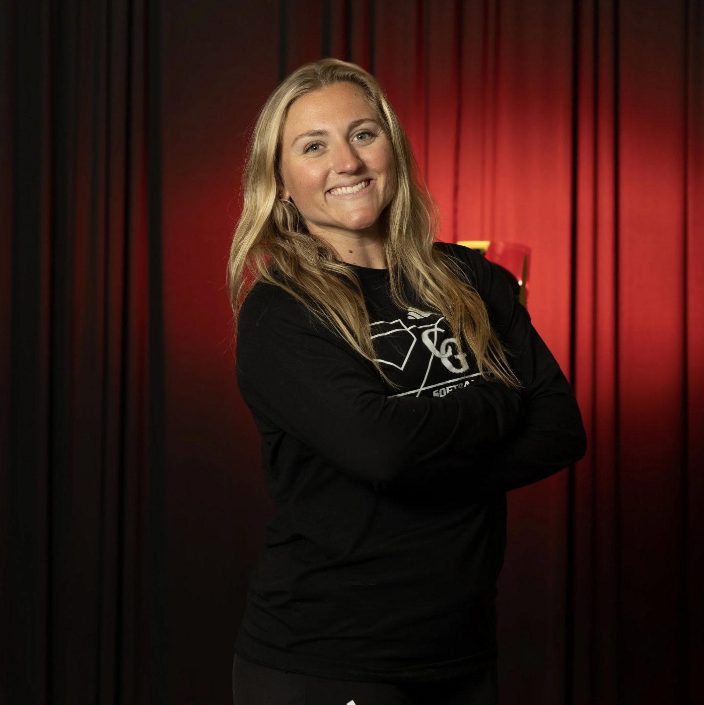 A young woman with long blonde hair smiling and crossing her arms, standing in front of red curtains.