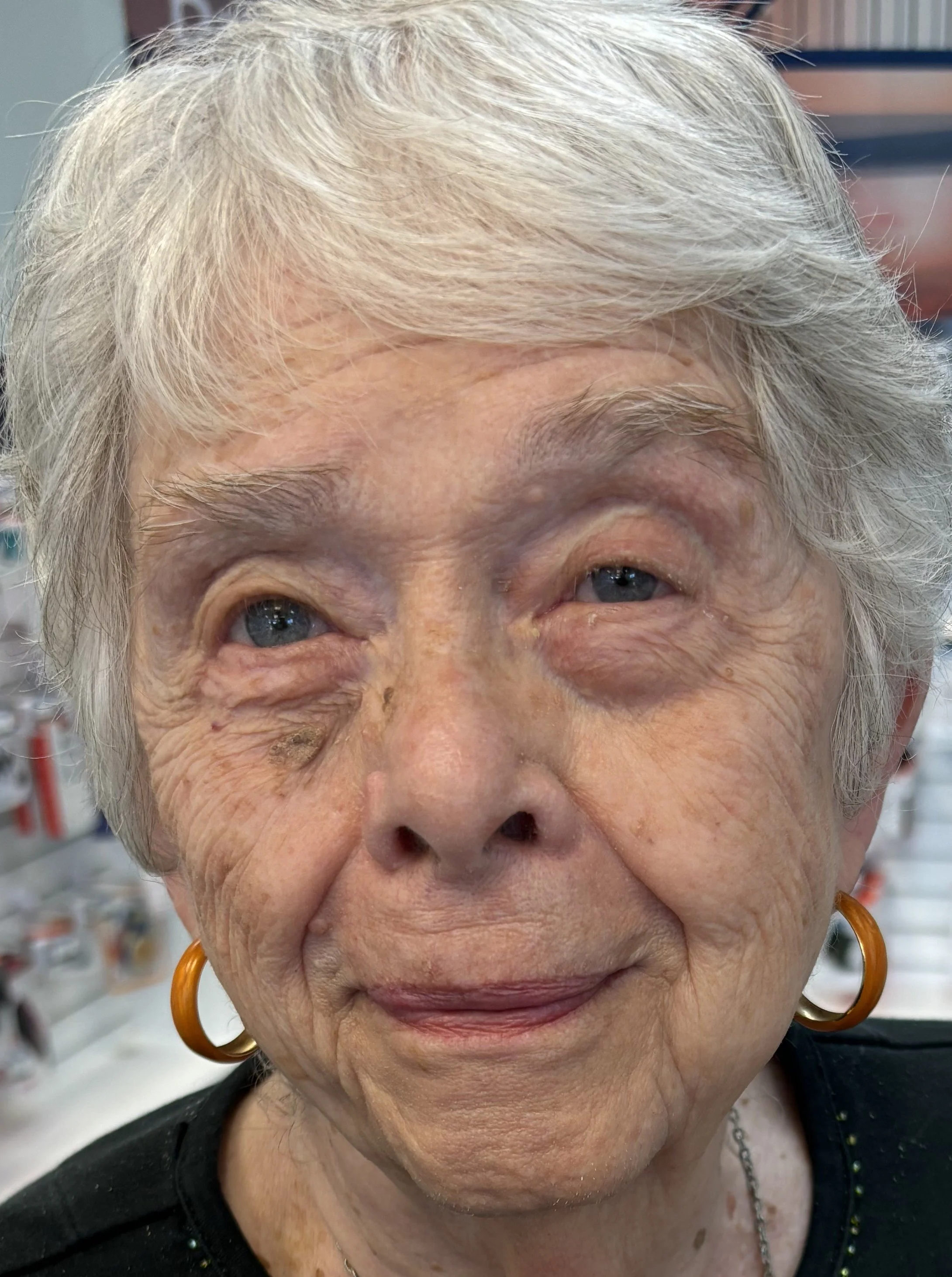 A close-up of an elderly woman with short white hair, wearing orange hoop earrings, smiling slightly, in a store with shelves of products in the background.