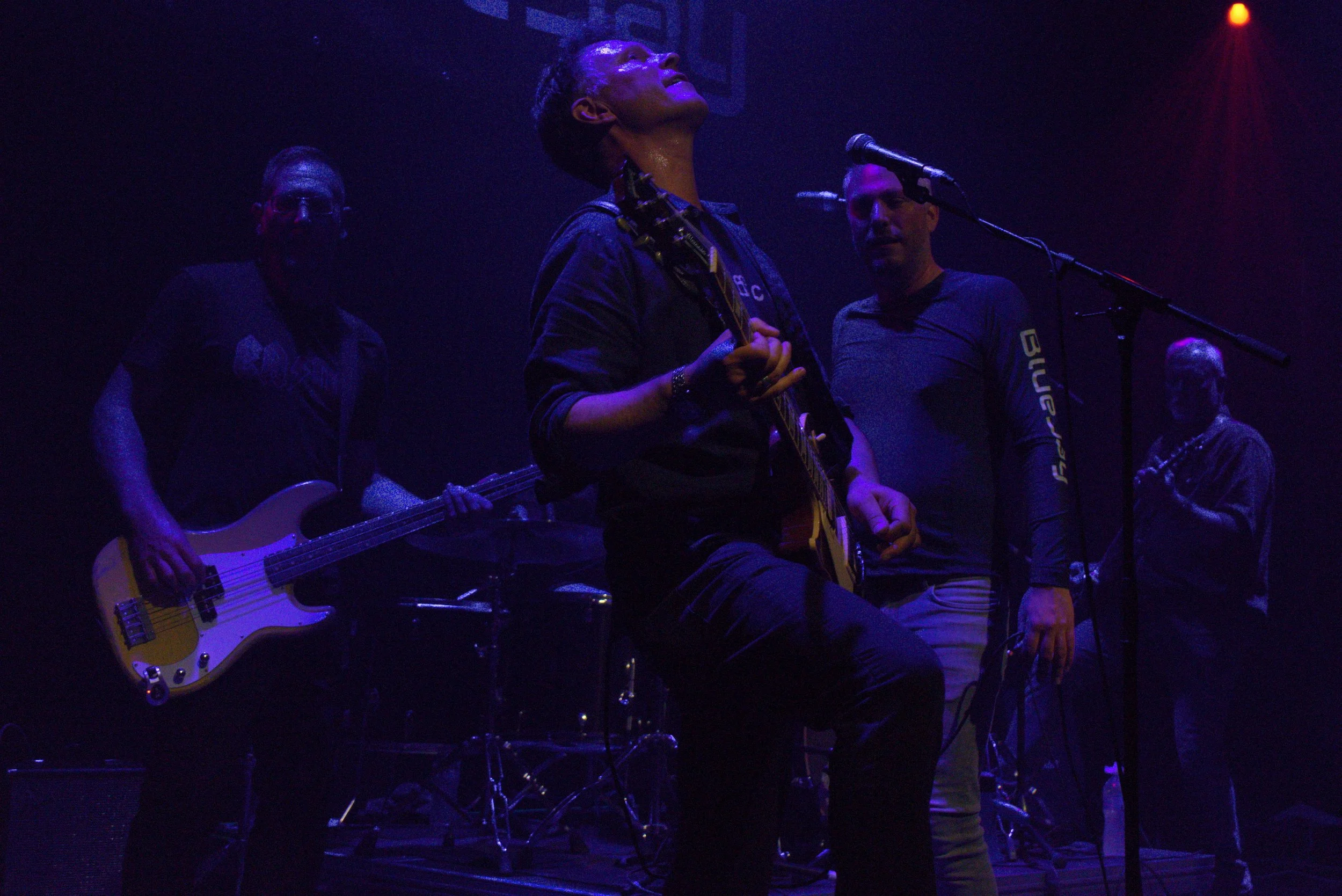 A live music performance on stage with five musicians. One playing bass guitar, another holding an acoustic guitar and singing, and others playing instruments in the background. Stage lighting is dark with purple and red hues.
