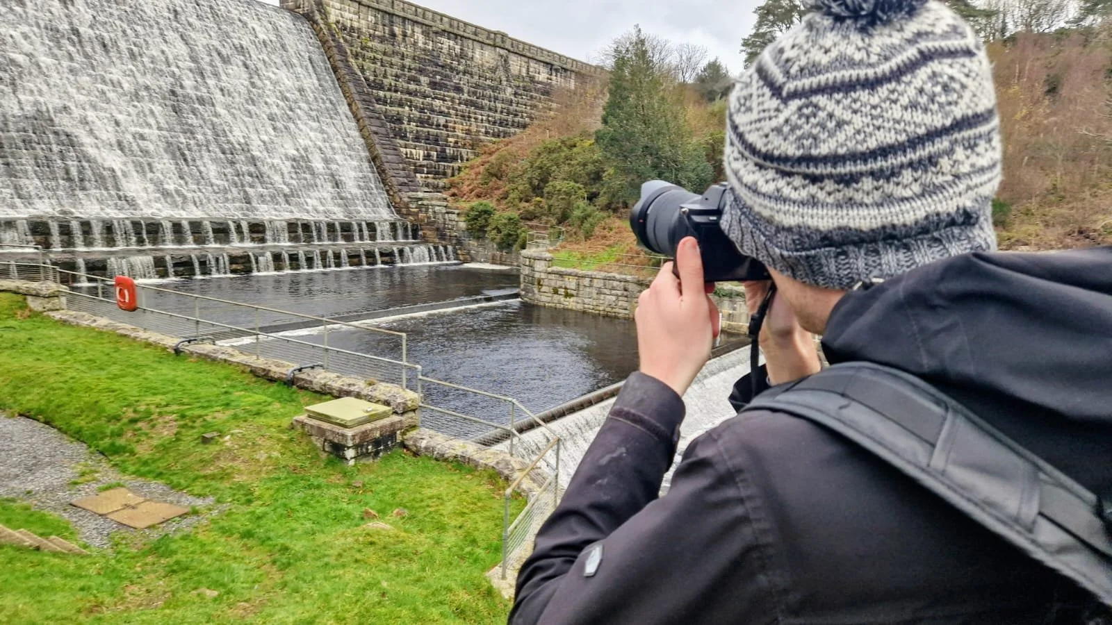 Person wearing a beanie and black jacket taking a photograph of a dam with a waterfall, water flowing over the spillway, surrounded by greenery and trees.