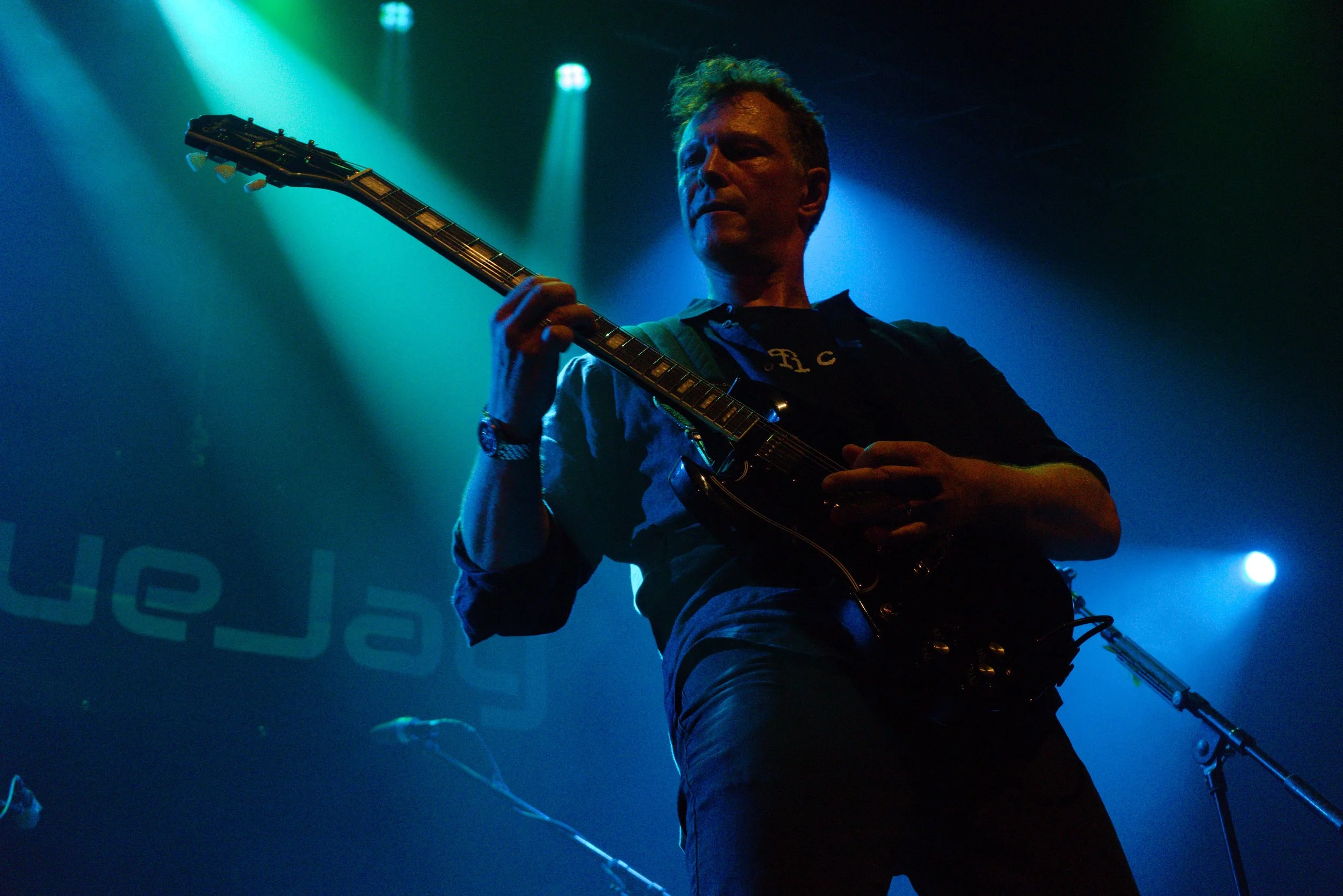 A man playing an electric guitar on stage with blue and green lighting, partially obscured by shadows.