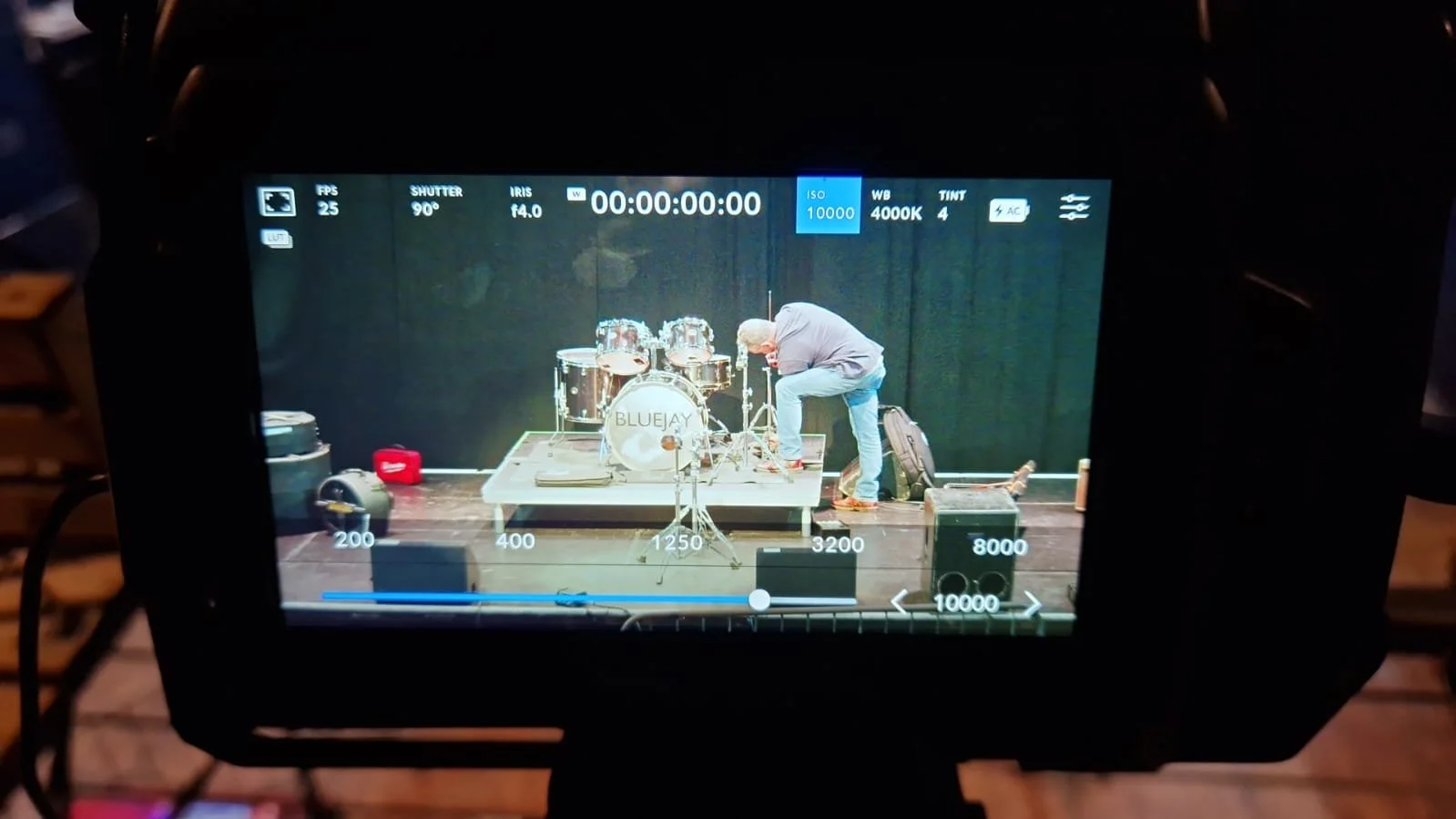 View of a camera screen showing a man adjusting a drum set labeled 'BLUEJAY' on a stage with percussion instruments and equipment around.