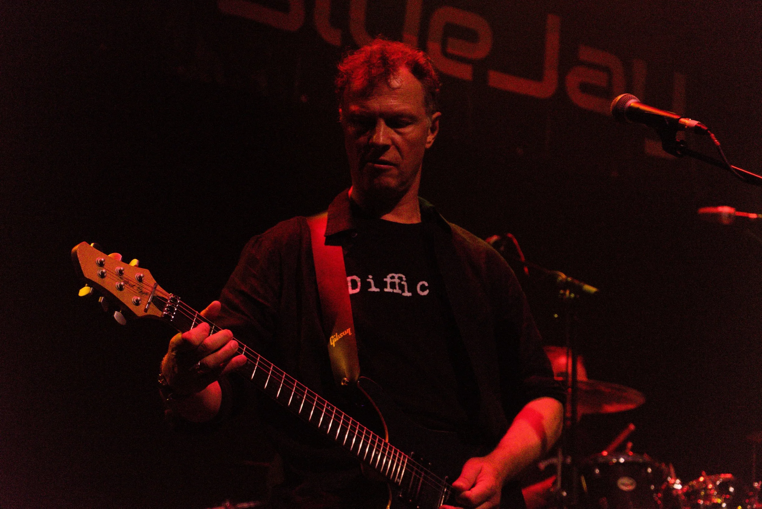 A man playing an electric guitar on stage with microphones and drums in the background, dimly lit with red lighting.