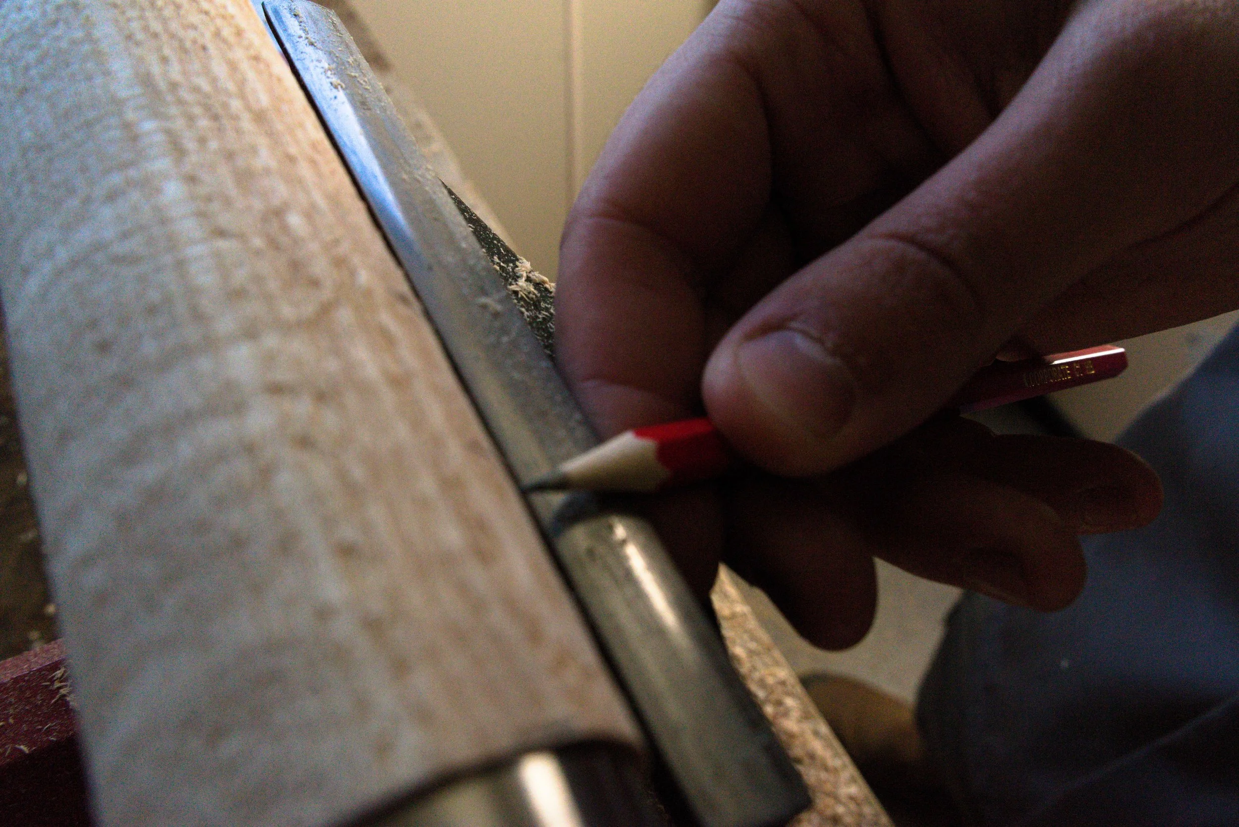 Close-up of a hand holding a red and white pencil, guiding it along a metal ruler on a wooden work surface, possibly marking or cutting a piece of material.