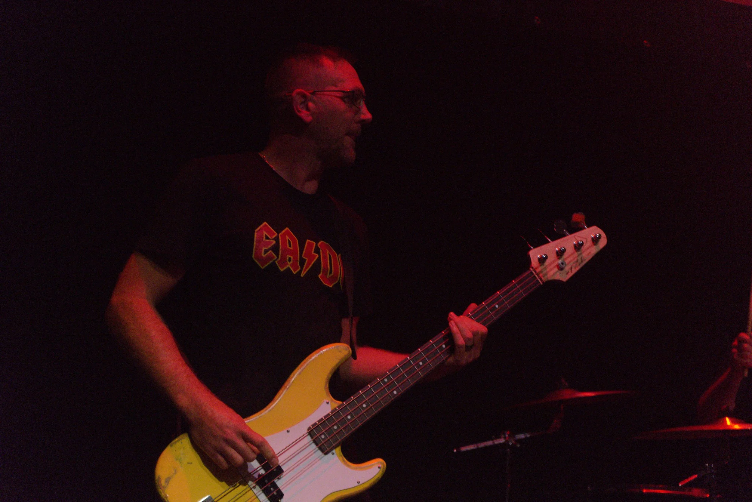 A man playing a yellow bass guitar on stage, illuminated by red stage lights, wearing glasses and a black T-shirt with red and yellow lettering.