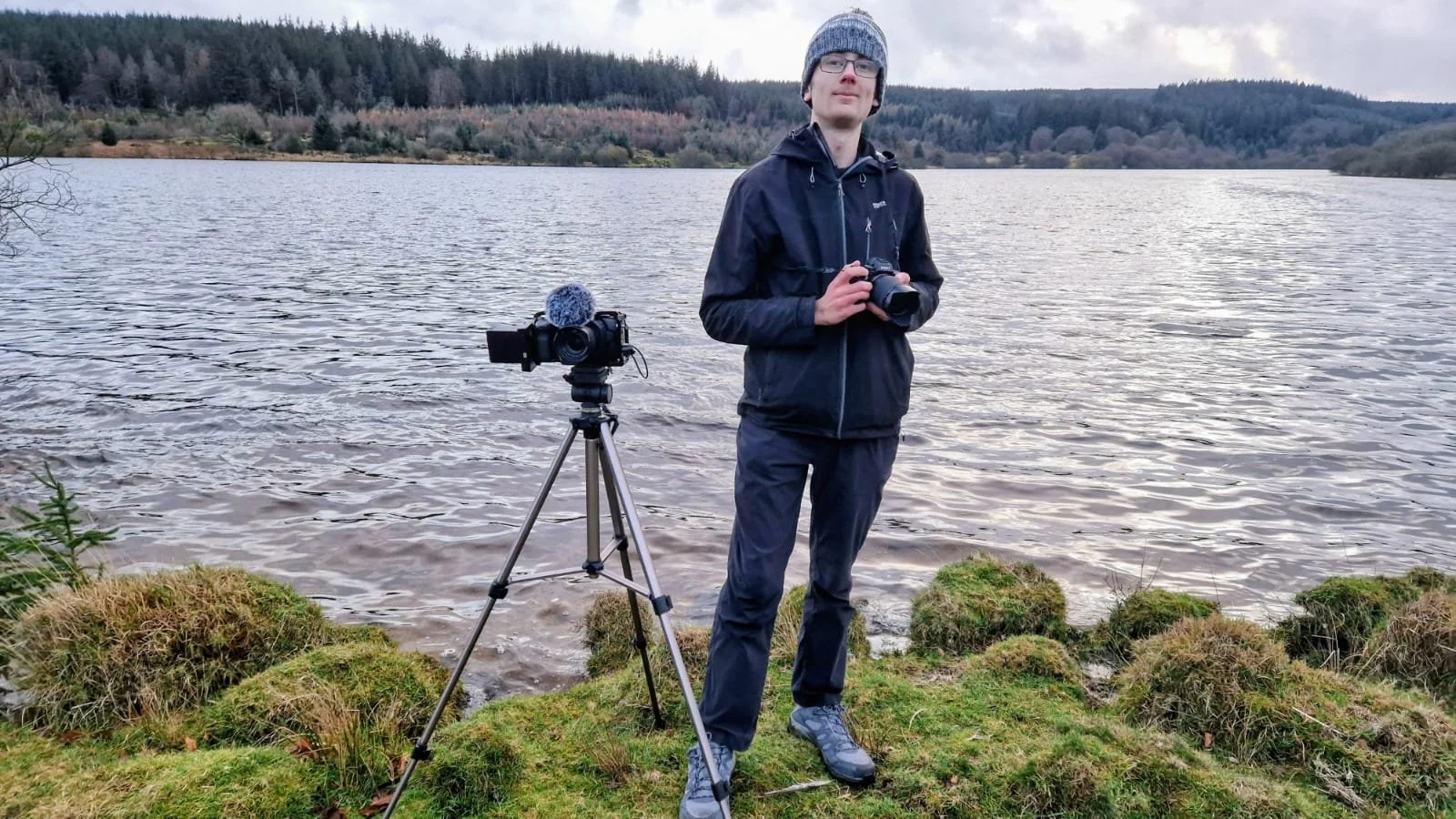 A man standing near a lake with a camera on a tripod and another camera in his hands, wearing a gray beanie, glasses, a black jacket, gray pants, and hiking boots, surrounded by mossy rocks and trees in the background.