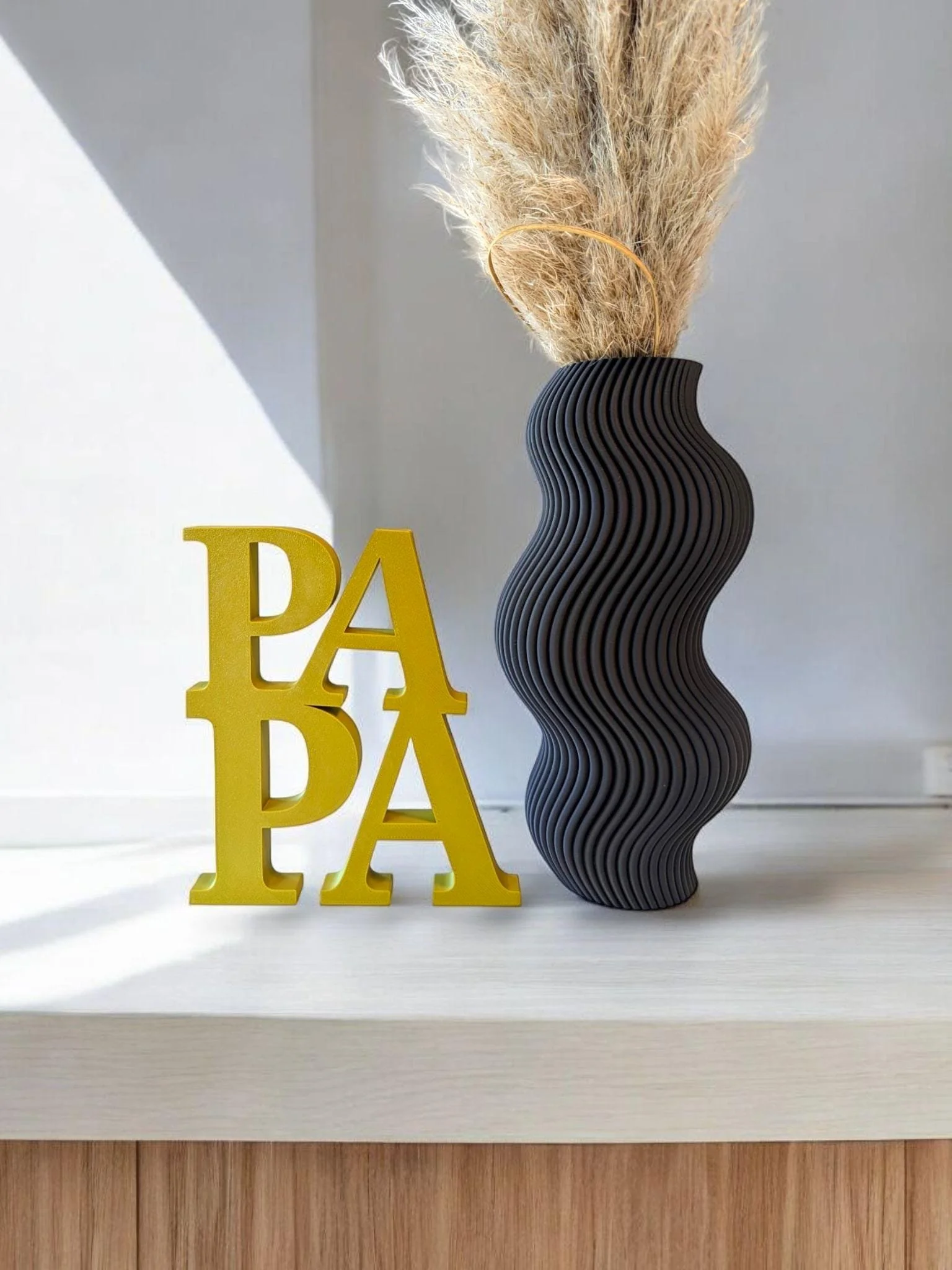 Eine schwarz-wellenförmige Vase mit getrocknetem Gräsern und der gelben Dekoration 'PAPA' auf einer weißen Oberfläche vor einer hellgrauen Wand.