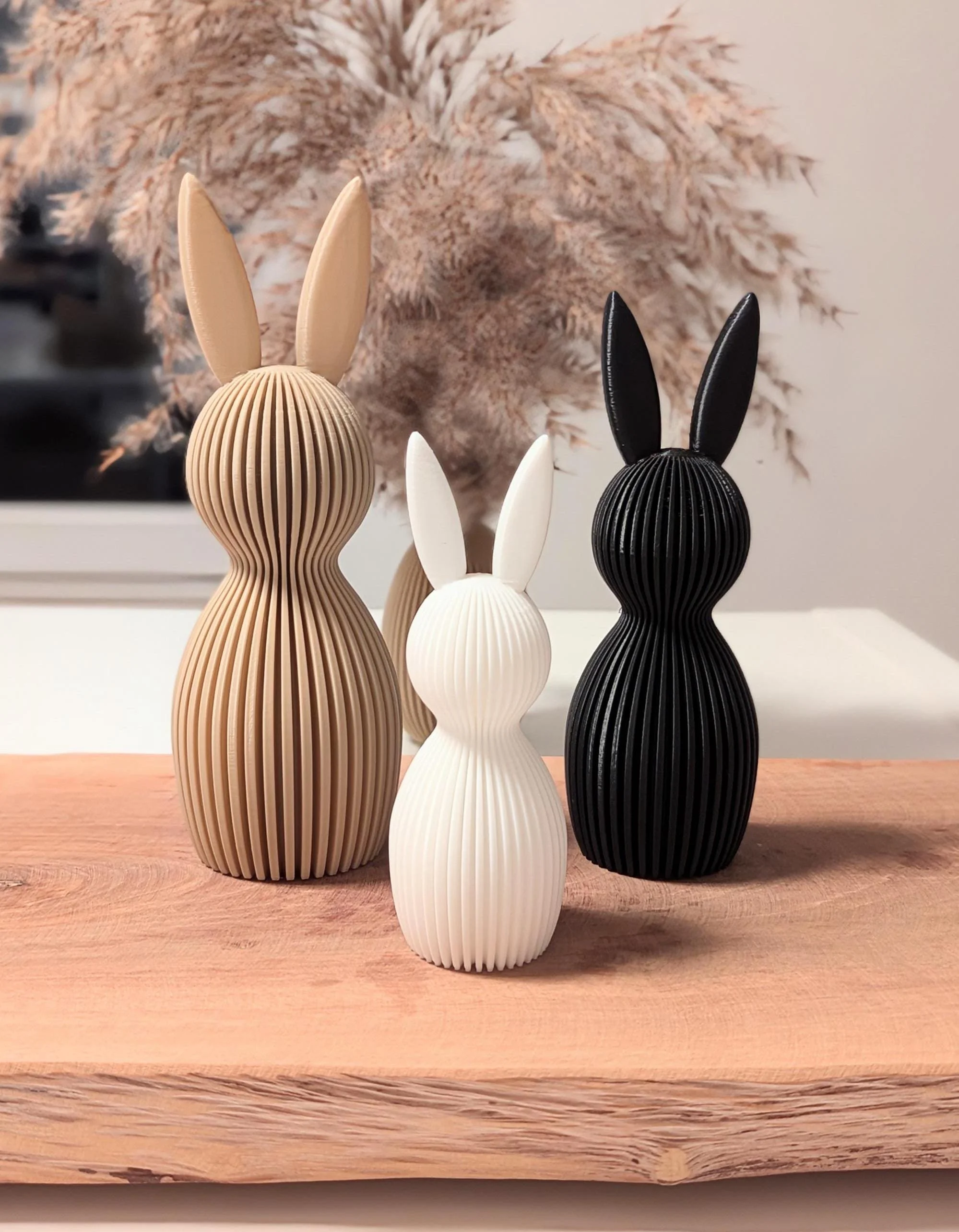 Drei stilisierte Hasenfiguren aus geripptem Material in beige, weiß und schwarz auf einem Holztisch, im Hintergrund besondere Trockenpflanzen.