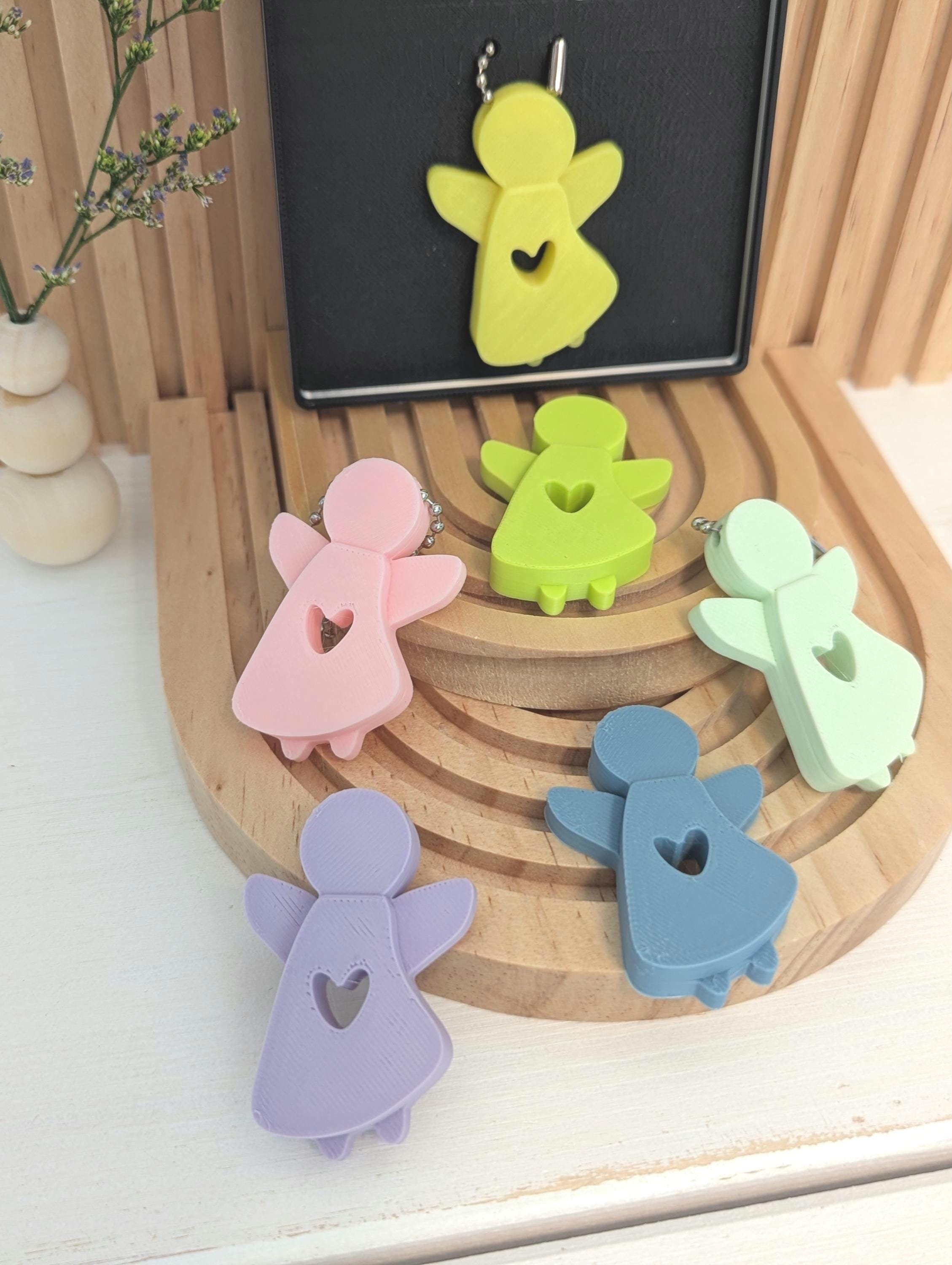 Bunte Schlüsselanhänger in Form von Engeln, aus Holz oder Kunststoff, zeigen Herzschlitz, auf einer runden Holzablage, mit einer kleinen Vase mit Blumen im Hintergrund.