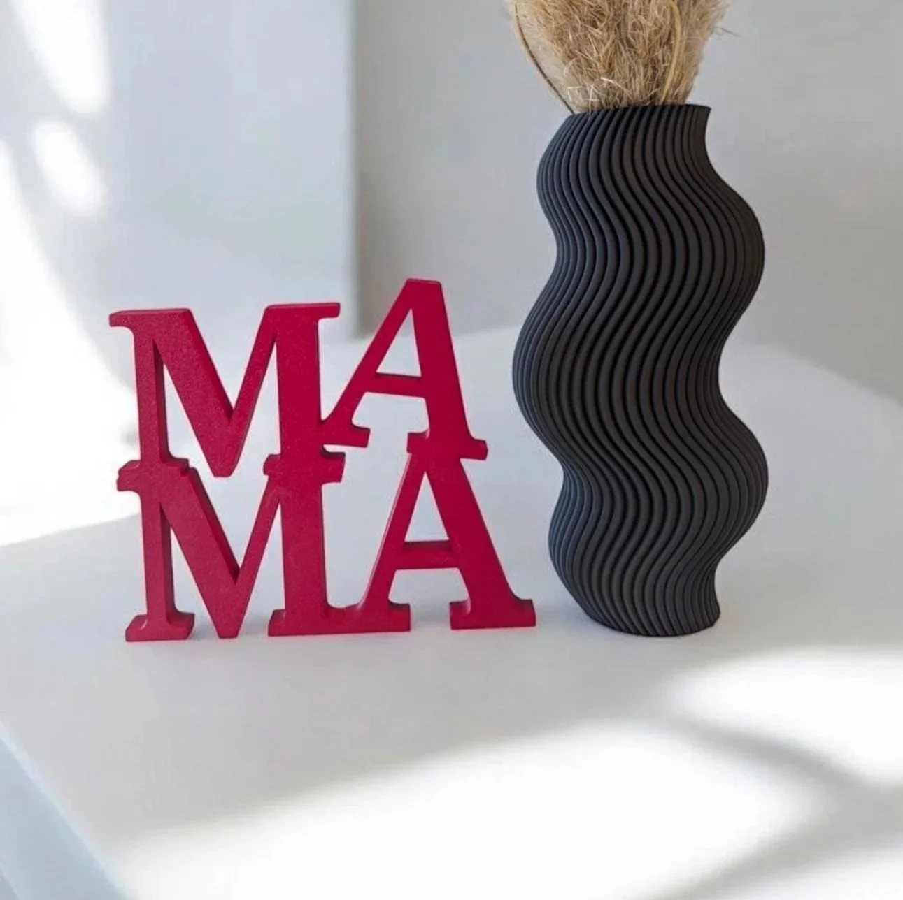 Eine rosa Deko mit the Buchstaben M und A, eine schwarze, gewellte Vase mit Holz als Inhalt.