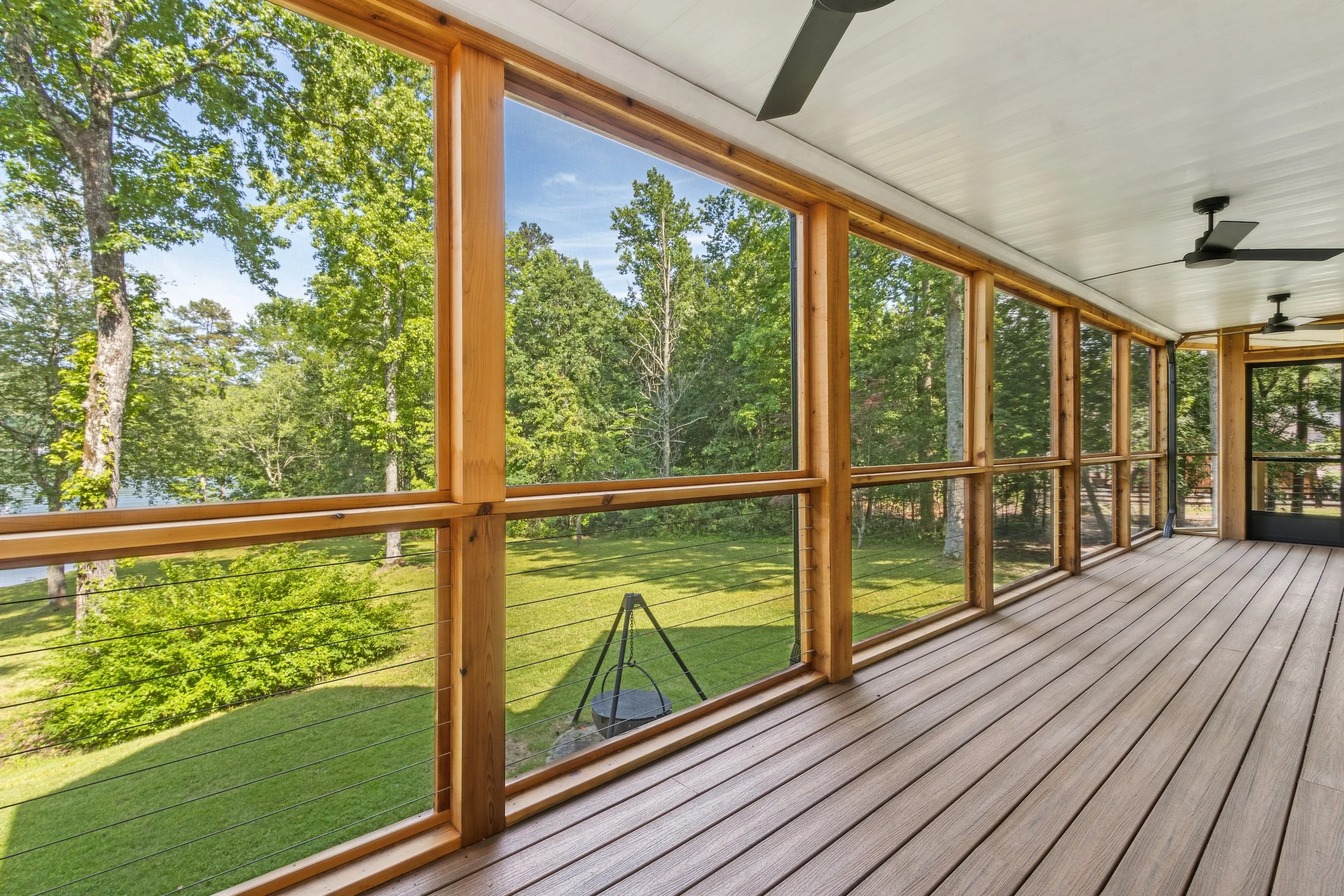 Covered porch with wooden flooring and large screened windows overlooking a green yard with trees and a lake.