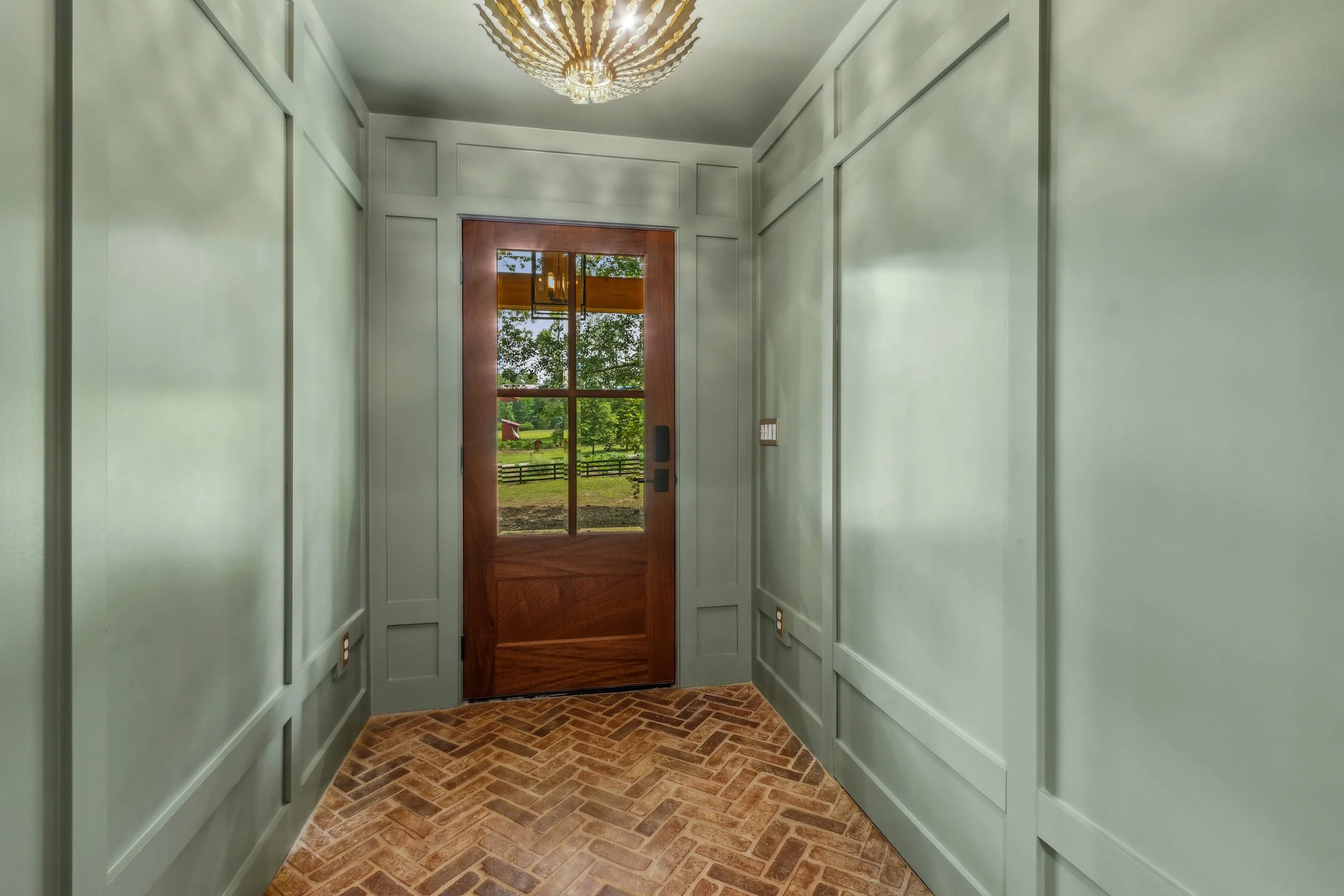 Entryway with a wooden door and large glass window, mint green paneled walls, brick herringbone floor, and a modern chandelier hanging from the ceiling.