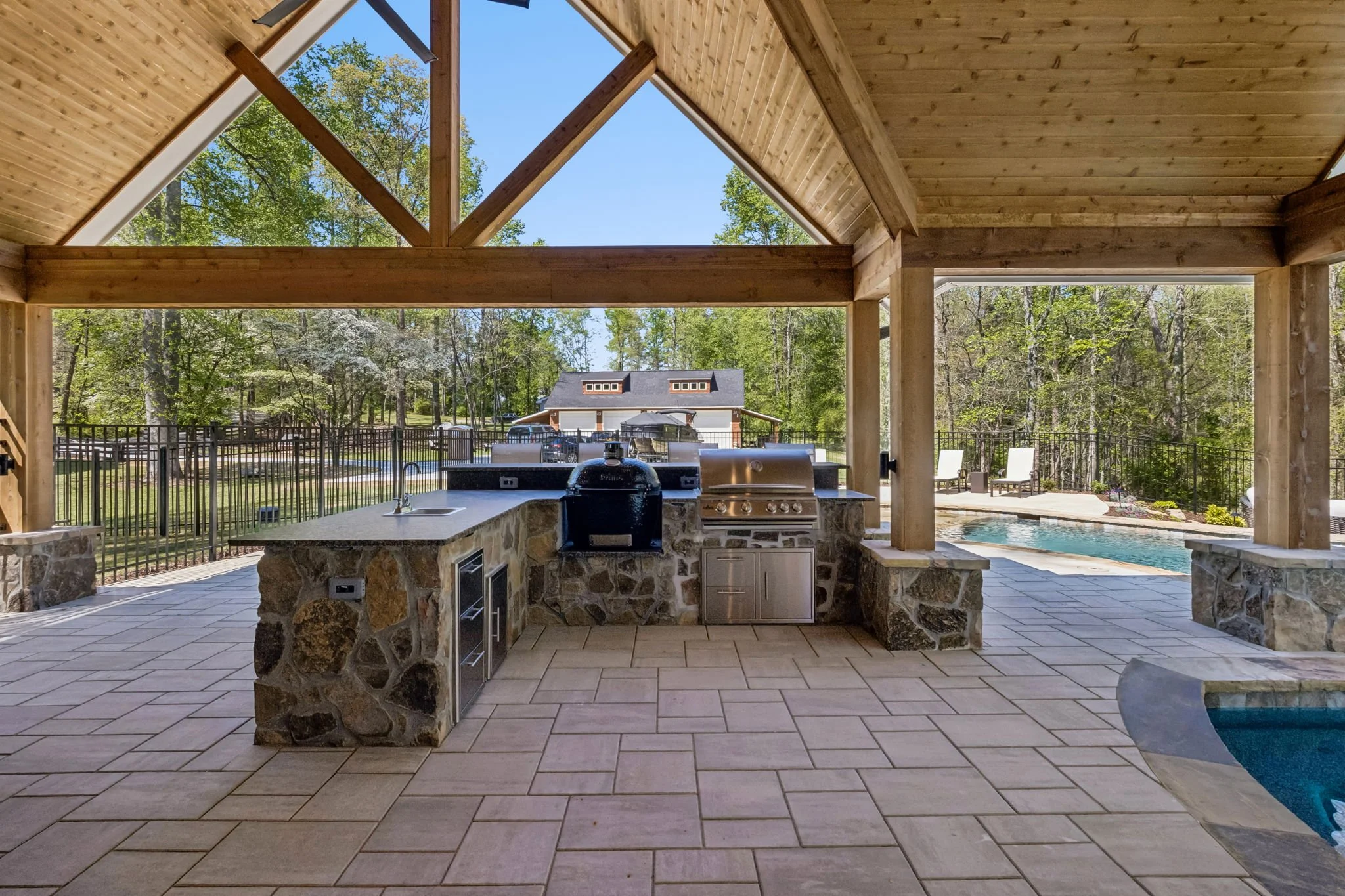 Royal Custom Homes & Remodeling created this backyard with a covered patio featuring a stone counter, built-in grill, a small sink, and a refrigerator, overlooking a fenced yard with a swimming pool, outdoor chairs, and a house in the background