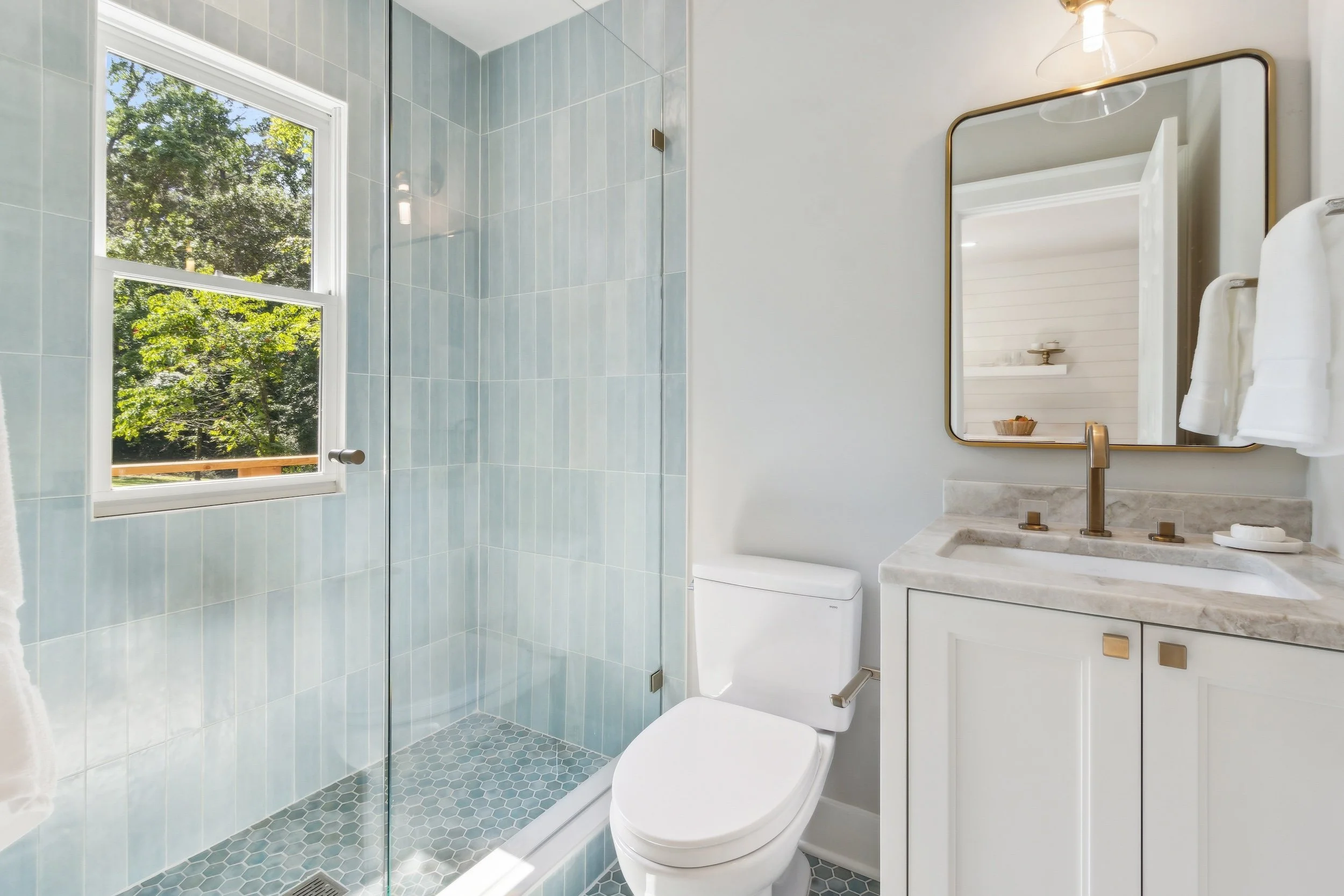 Bathroom with a walk-in shower, a window showing trees outside, a toilet, a white vanity with a marble countertop, a large mirror, and white towels hanging on the wall.