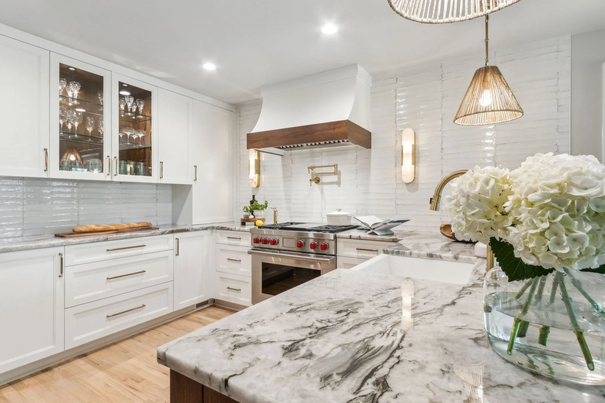 Kitchen counter with marble surface, a white farmhouse sink, a gold faucet, a wooden bowl of artichokes and a tomato, beige pendant lights, a vase of white flowers, and large windows showing green trees outside.