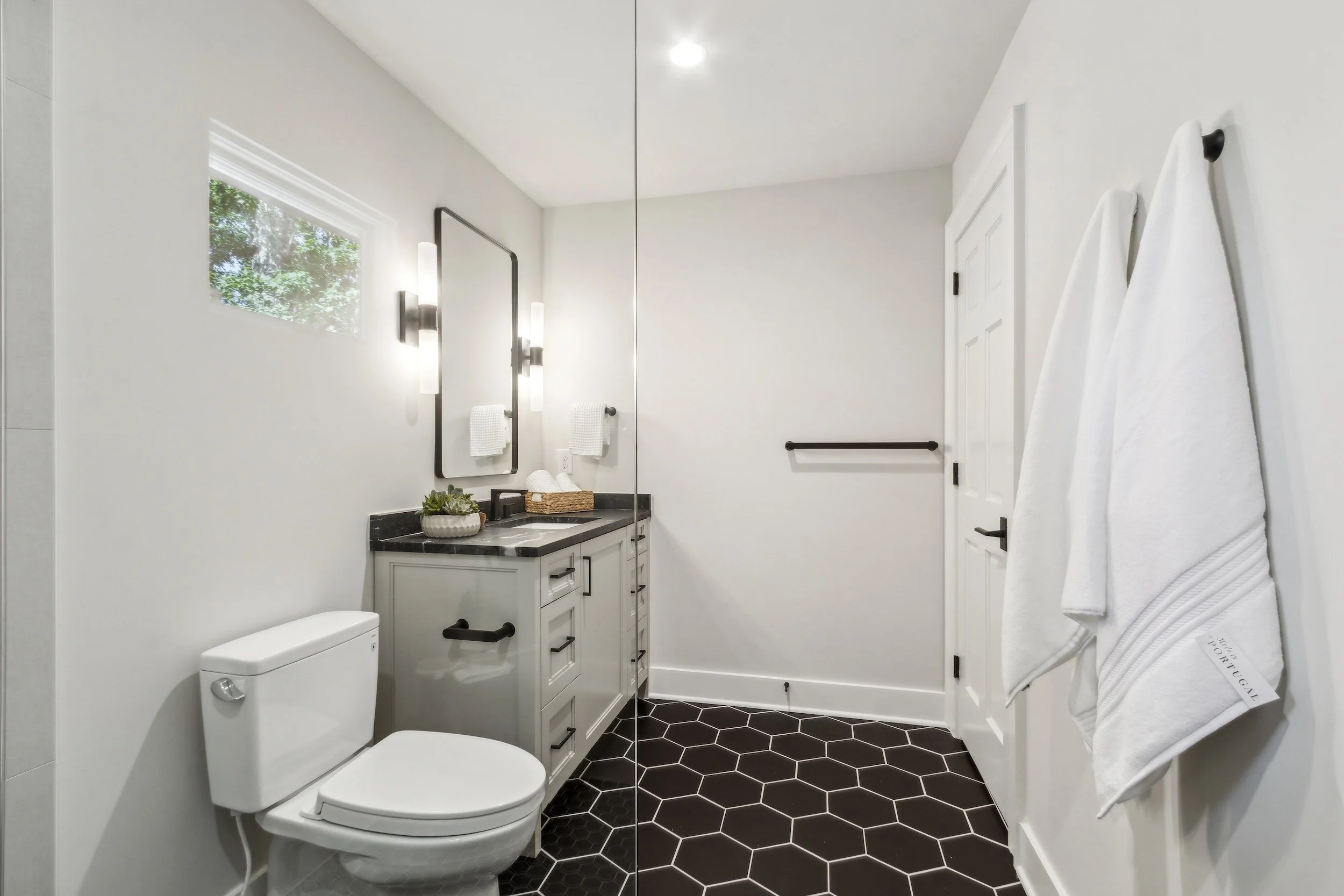 A modern bathroom with a white toilet, a gray vanity with a black countertop, a large mirror, wall-mounted light fixtures, a small window, white towels, and a black hexagonal tile floor.