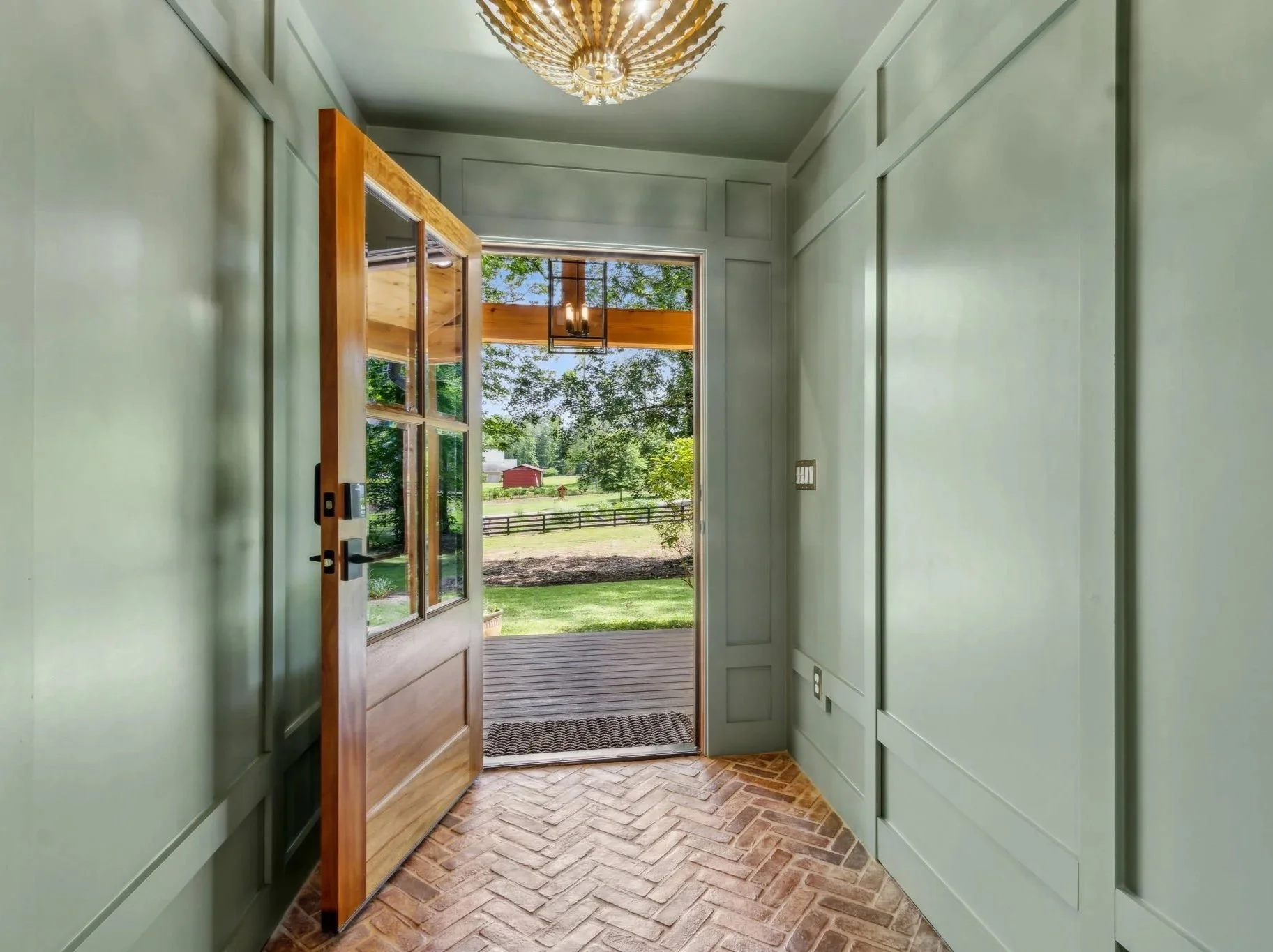 Interior entryway with a wooden door open to a porch and a view of green fields and trees outside, featuring light-colored walls and brick flooring.