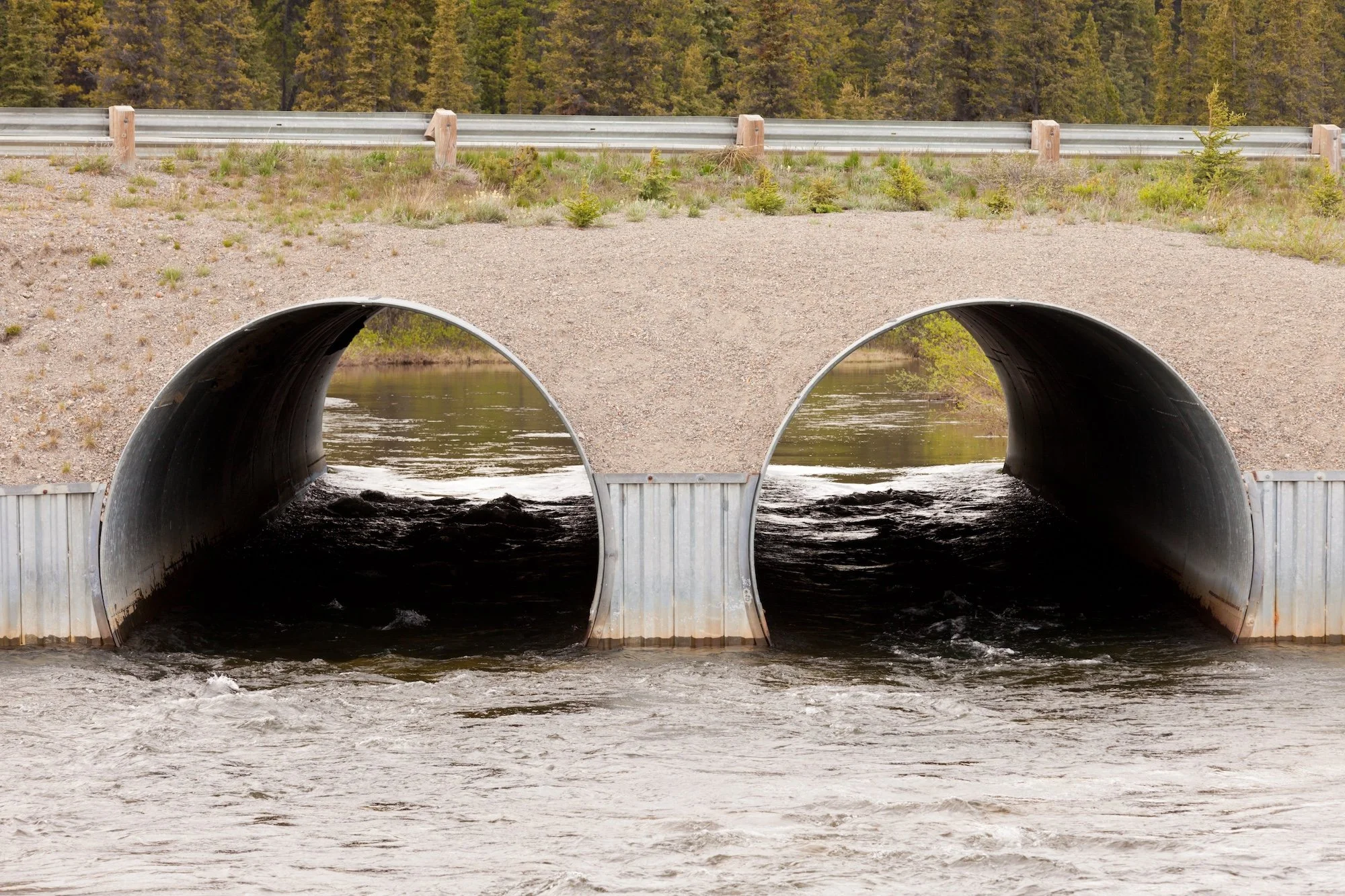 Two large metal culverts under a dirt road, allowing water to flow through, with water visible inside and flowing out at the bottom, surrounded by grass, trees, and a forested area in the background.