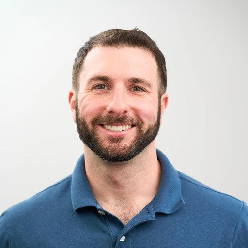 A smiling man with short dark hair, a beard, wearing a blue polo shirt, against a plain light background.