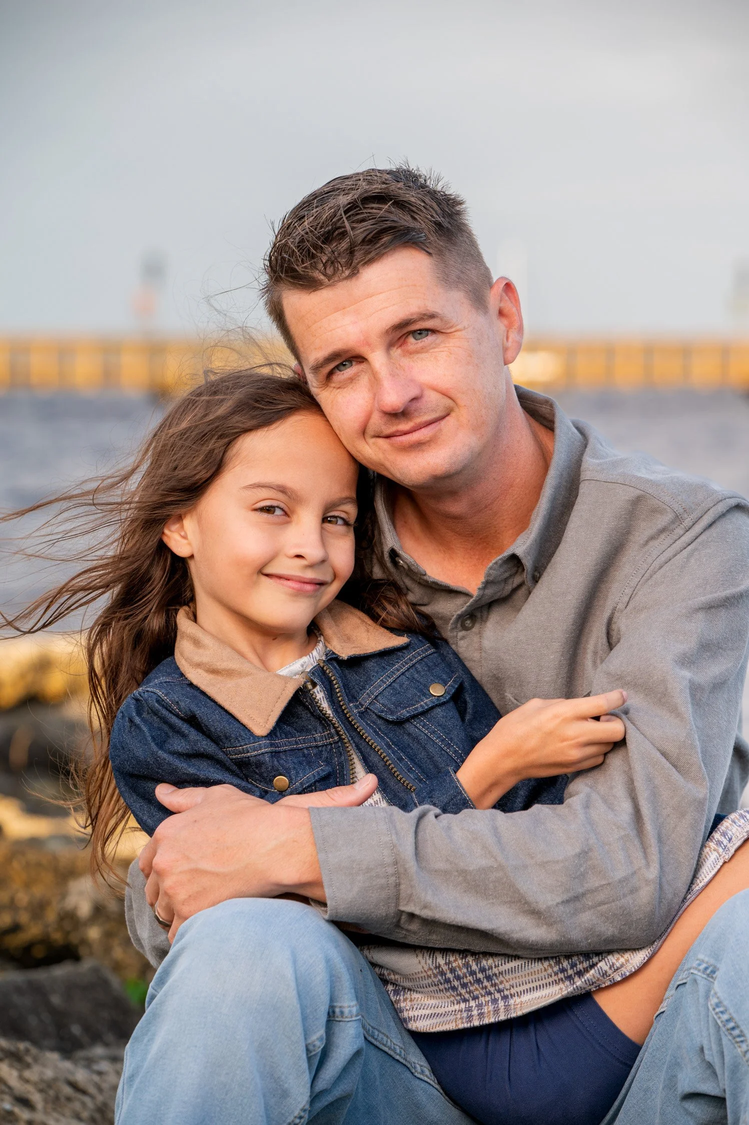 A man and a young girl hugging outdoors on a rocky surface with a blurred harbor or dock in the background.
